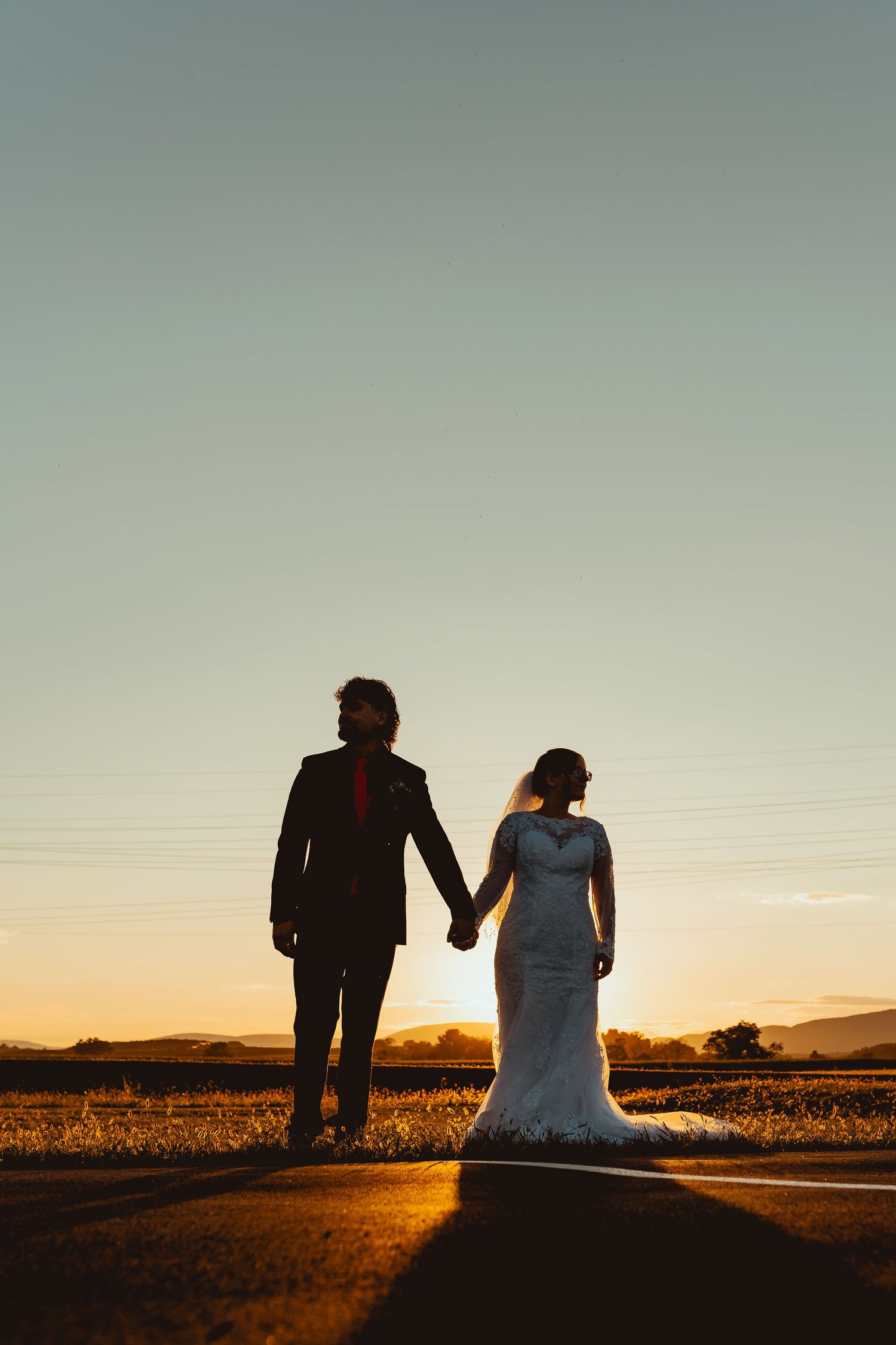 Silhouette of bride and groom holding hands at sunset on a road, golden hour wedding portrait.