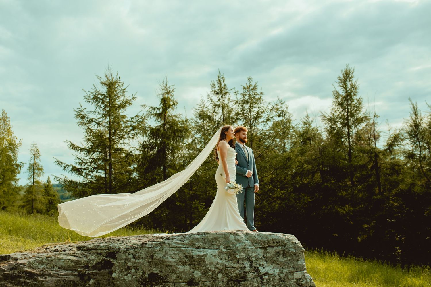 A wedding couple stands on a rocky overlook as the bride's long veil flows dramatically in the wind.
