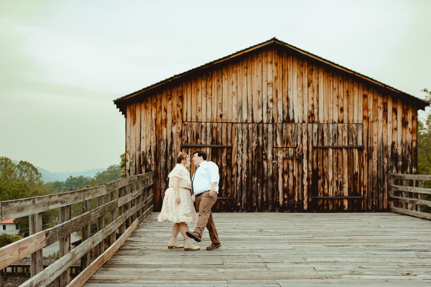 A couple stands together on a wooden boardwalk in front of a rustic barn on an overcast day.