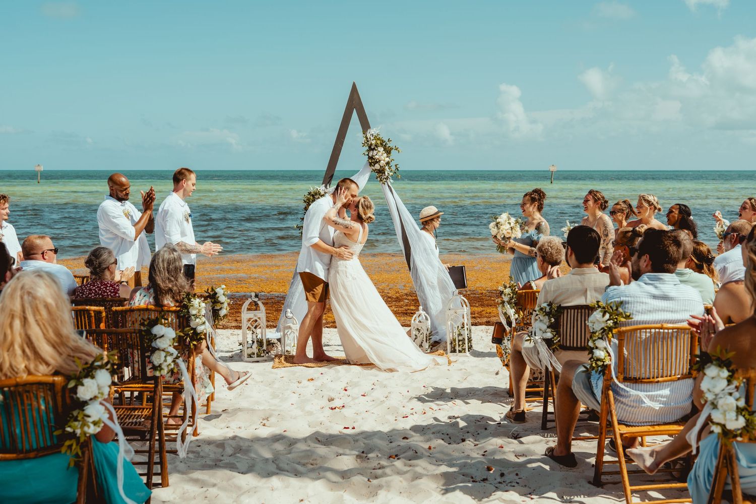 Couple kissing at a romantic beach wedding ceremony with a triangular arch, floral decorations, and guests seated on sandy shore.