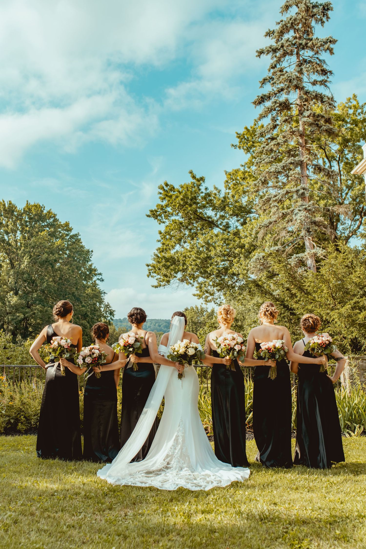 A wedding party in black dresses and white gown poses together on a grassy hill under blue skies.