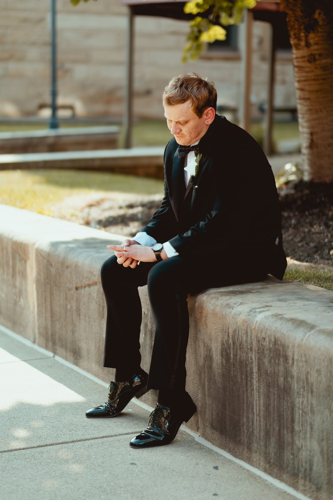 A figure in a black suit sits alone on concrete steps looking down at a mobile device.