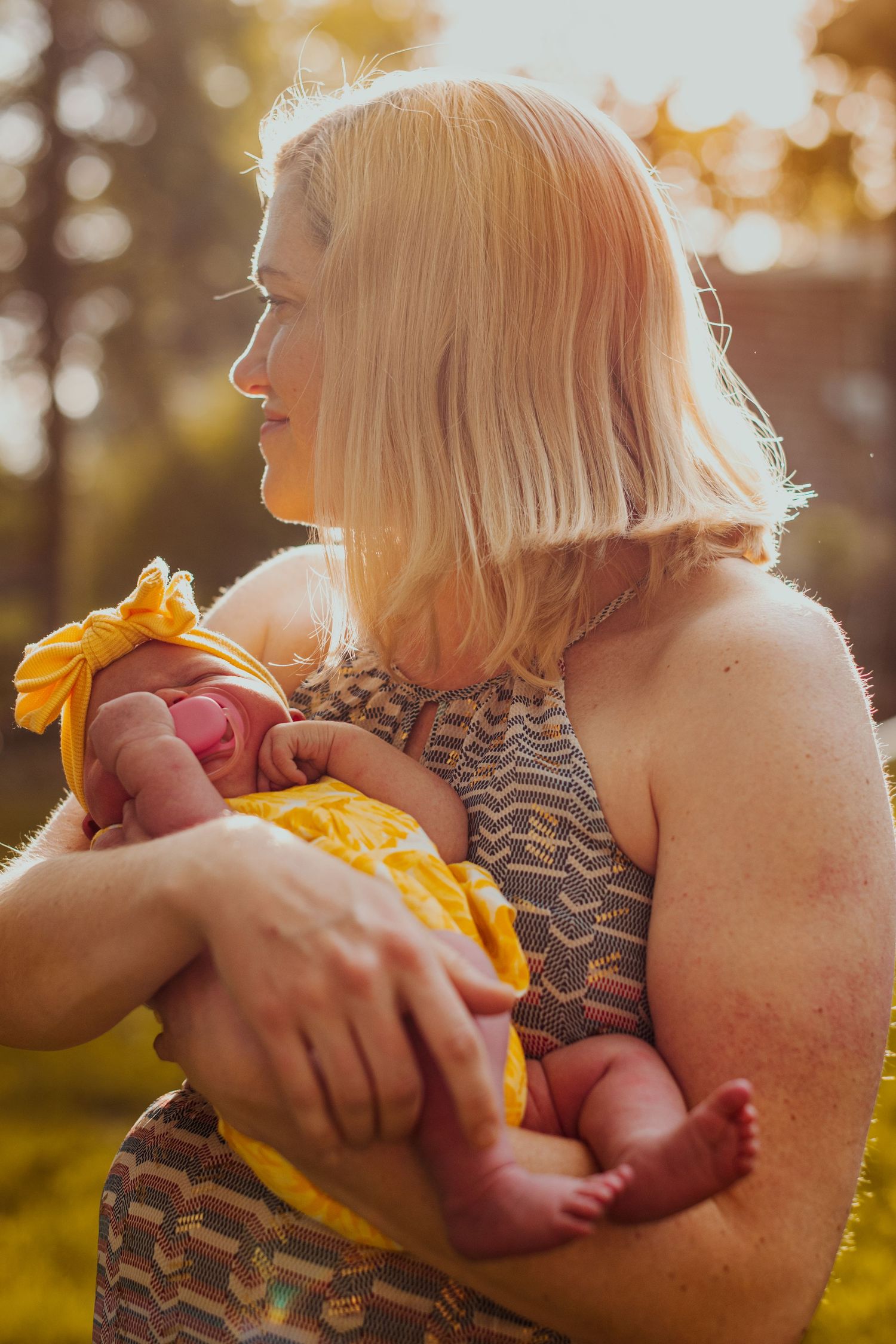 A tender outdoor photoshoot showing a parent cradling a newborn baby in golden sunlight.