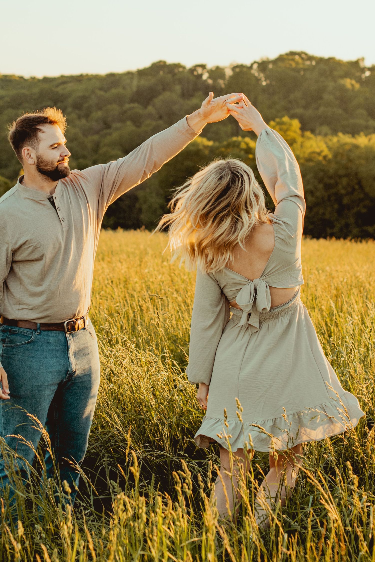 A couple twirls and dances through a sun-drenched field of tall grass during golden hour.