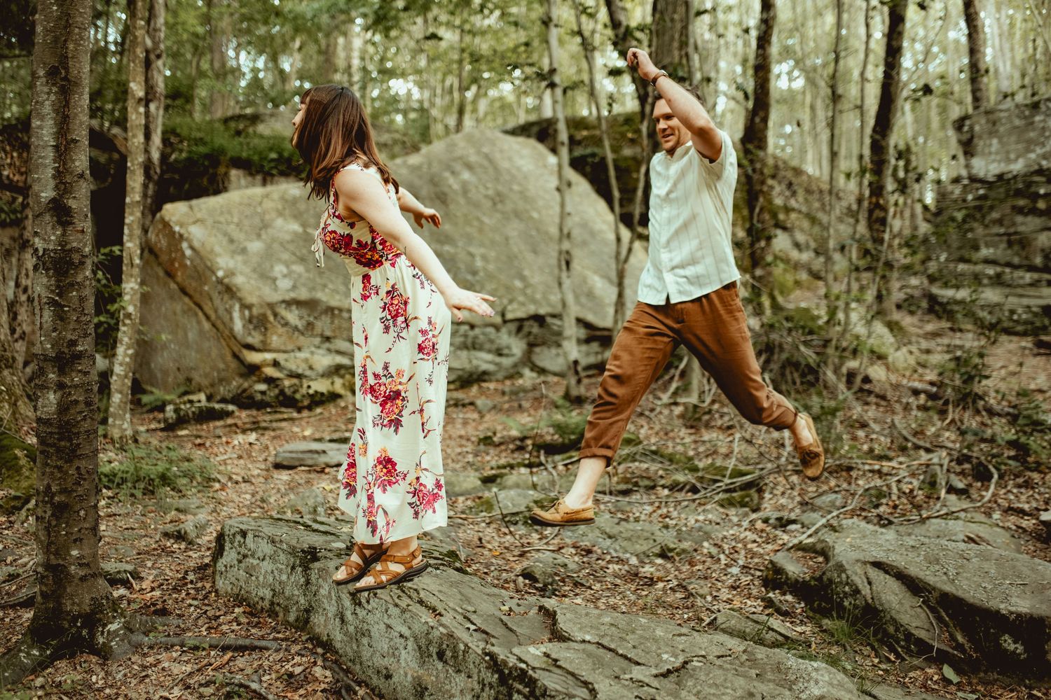 Couple dancing on rocks in a forest, woman in floral dress holding hands with man in brown pants.