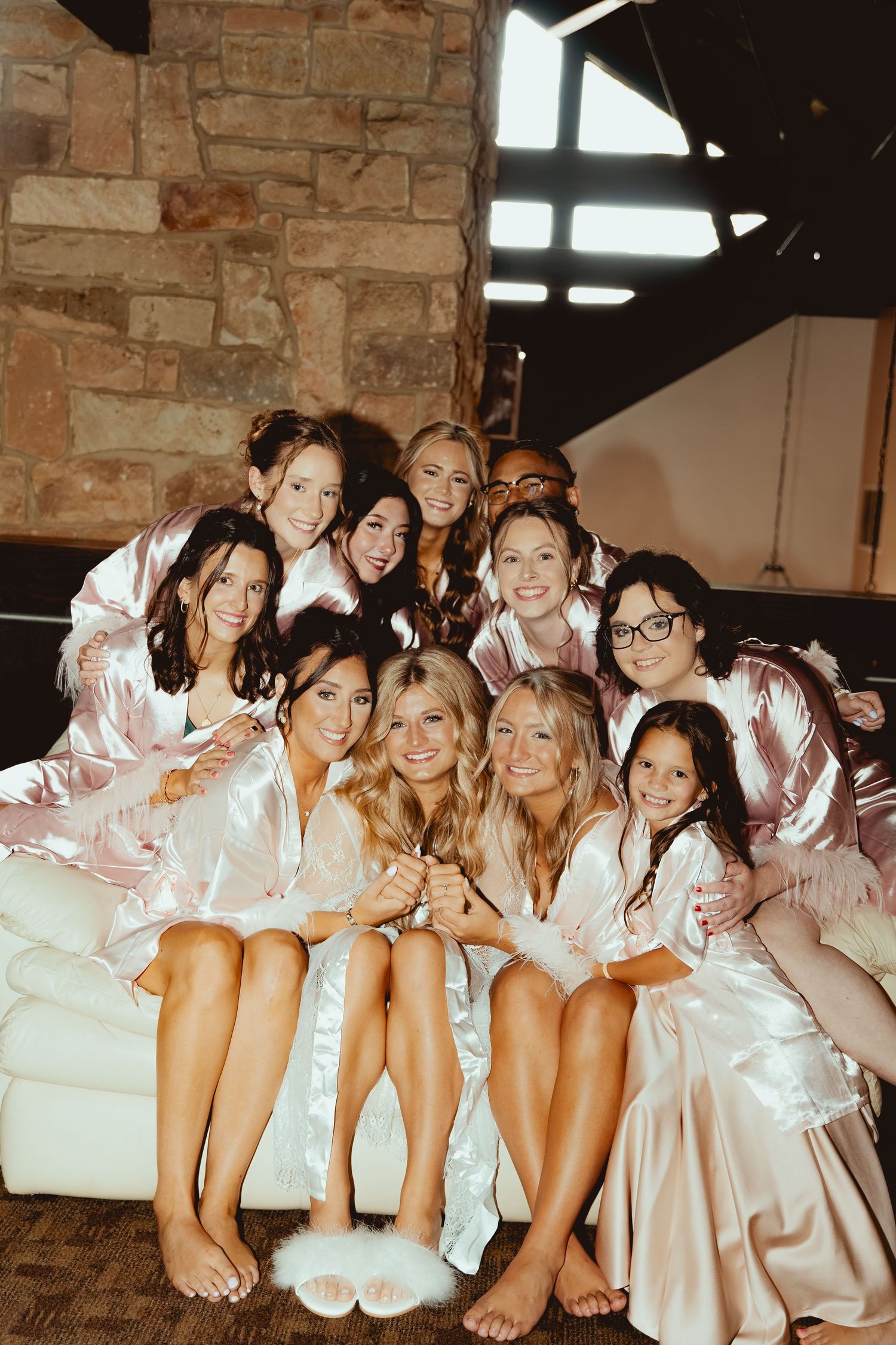 Bride and bridesmaids wearing matching satin robes pose together on a couch near a stone fireplace.