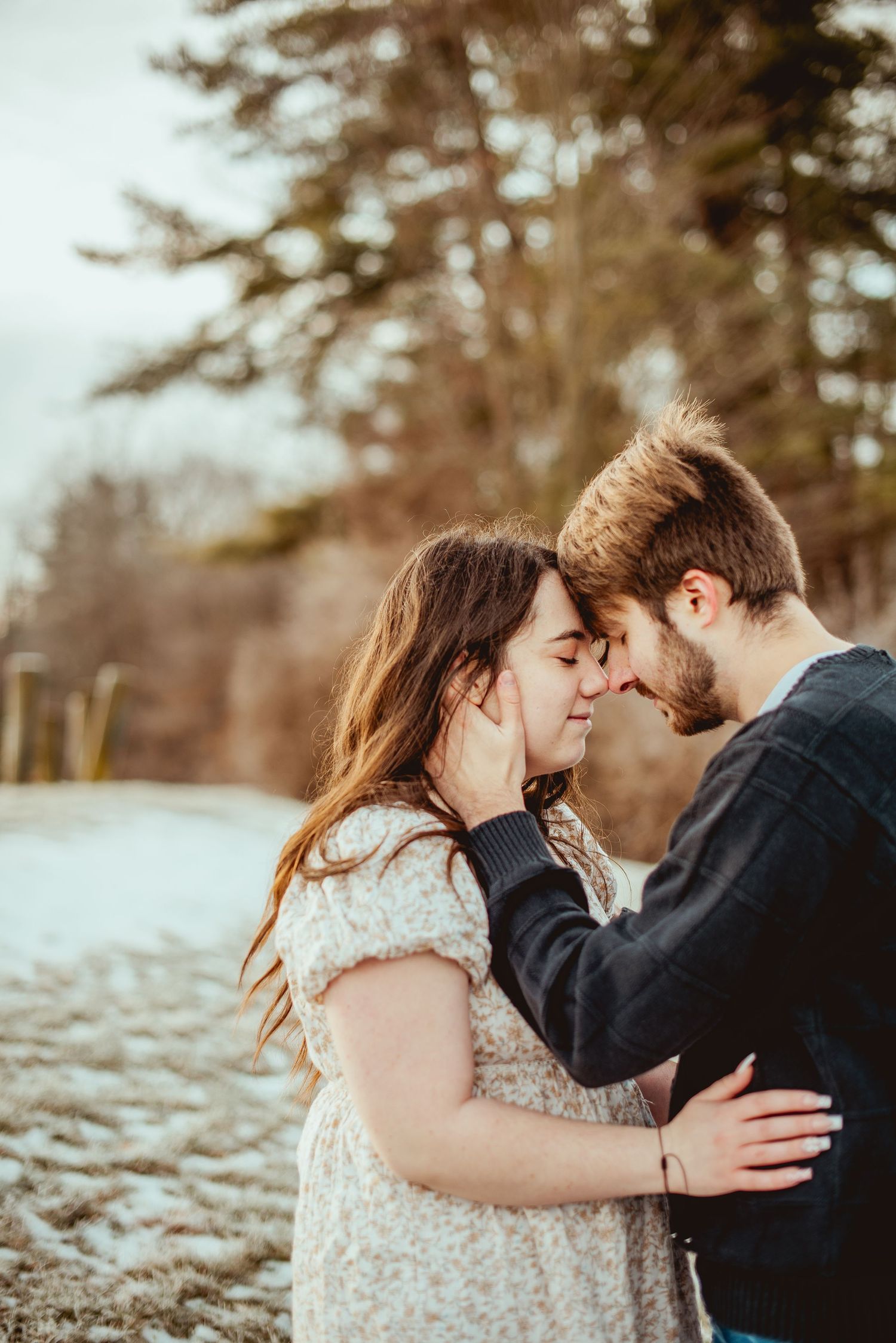 A romantic moment captured between a couple in winter with snowy landscape and evergreen trees behind them.