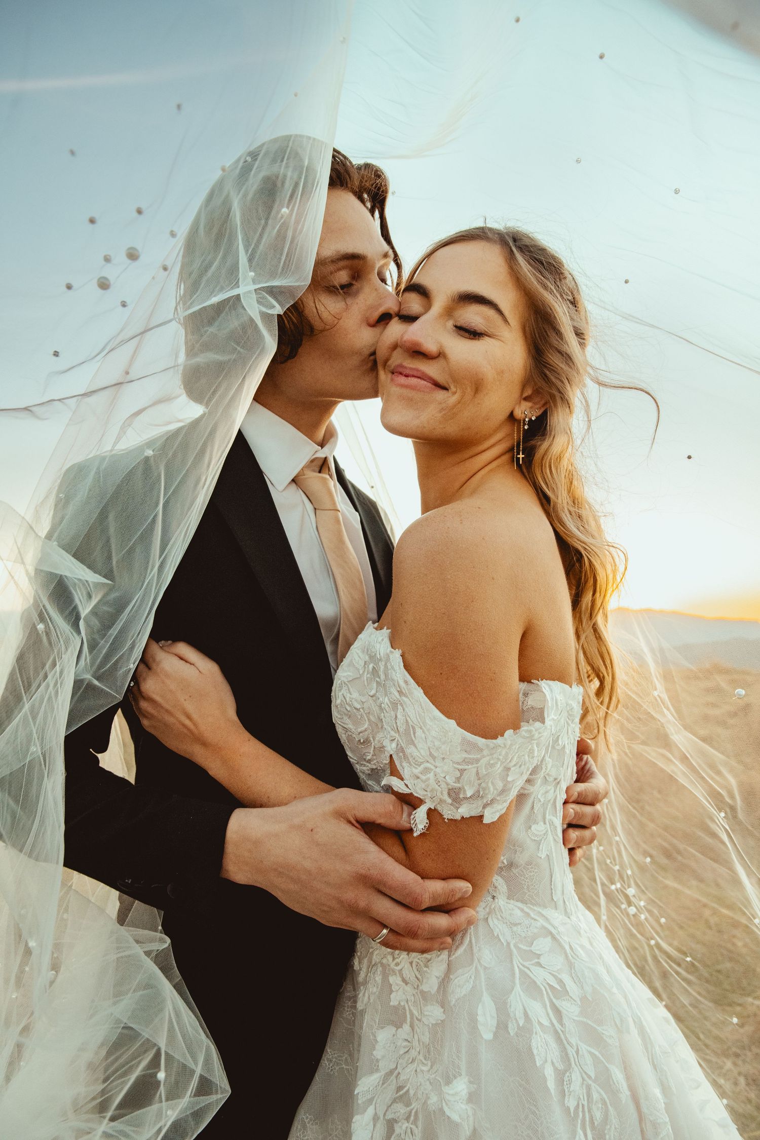 Intimate wedding portrait captured in warm sunlight with a flowing veil and mountain backdrop.