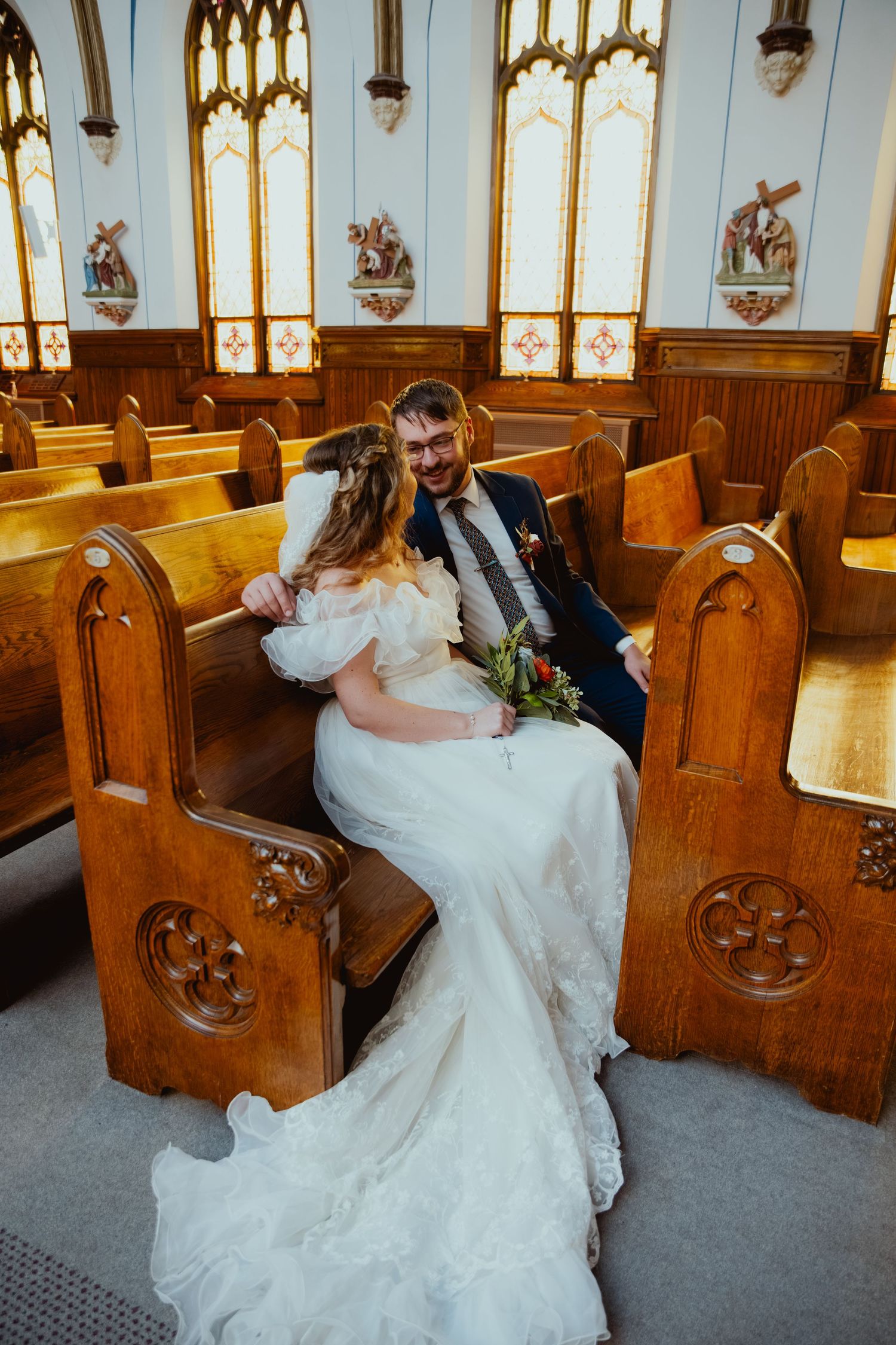 Bride in a white lace wedding gown sits with her groom holding a bouquet on a wooden church pew beneath stained glass windows.