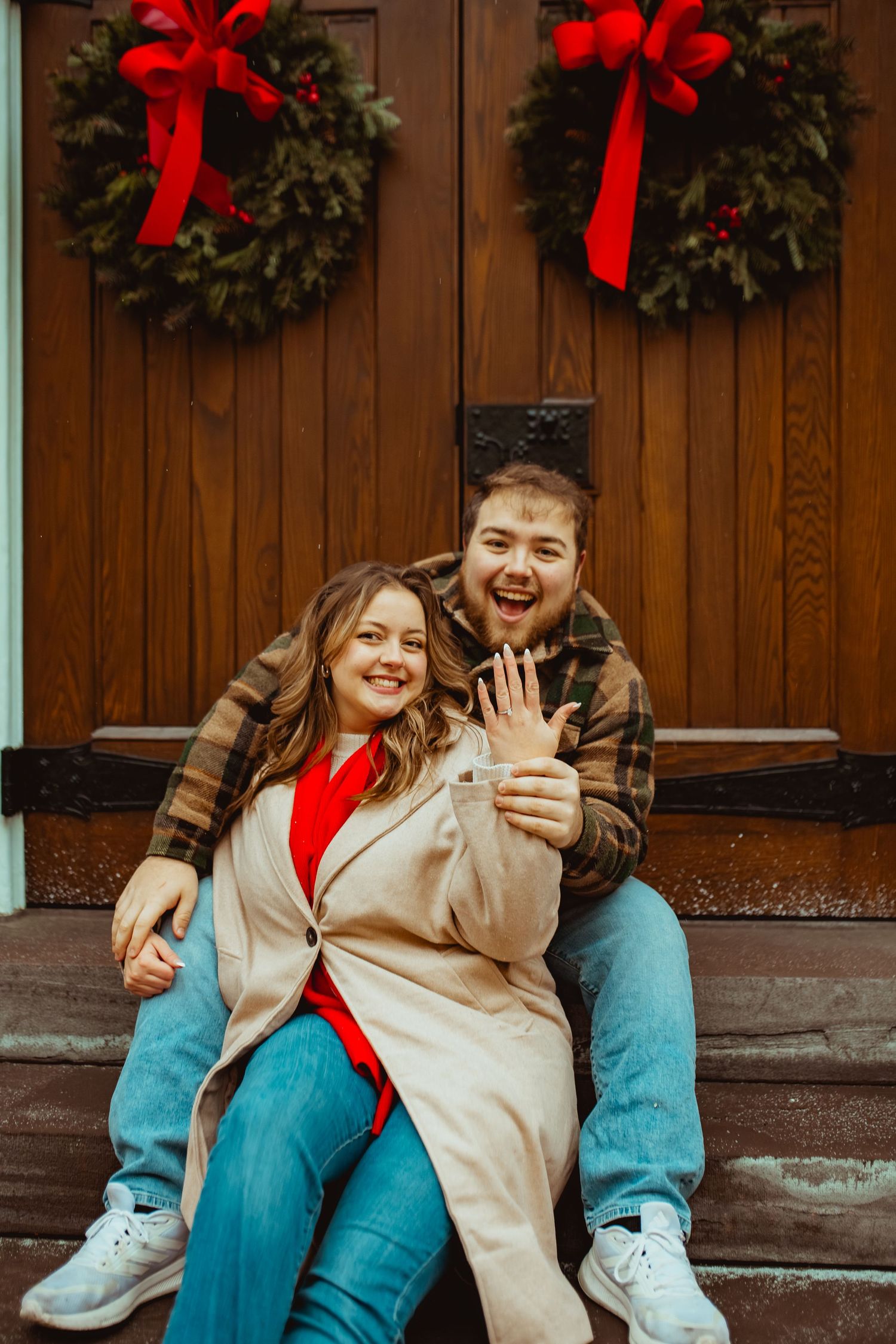 A couple laughs together on wooden steps decorated with holiday wreaths and red bows.