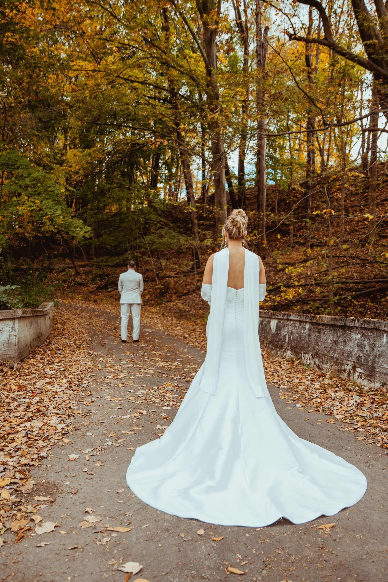 A wedding dress with a flowing train displayed in a fall forest setting with scattered autumn leaves on the ground.