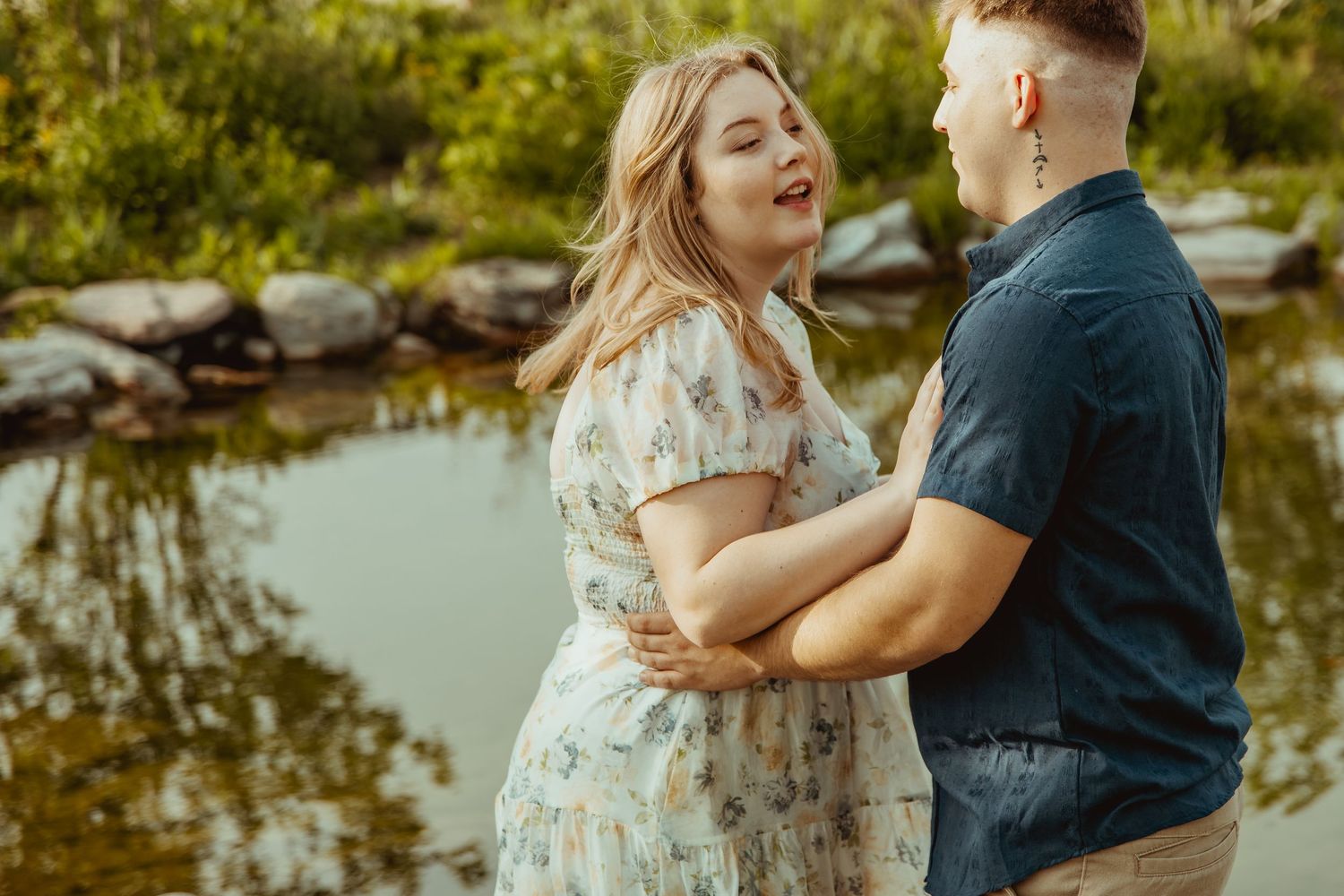 Couple sharing a romantic moment by a serene pond, woman in floral dress laughing joyfully outdoors.