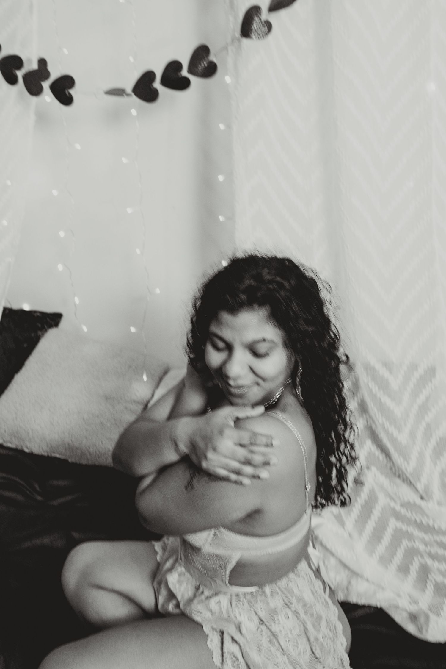 Black and white photo of a woman with curly hair sitting on a bed, hugging herself near heart garland decorations.