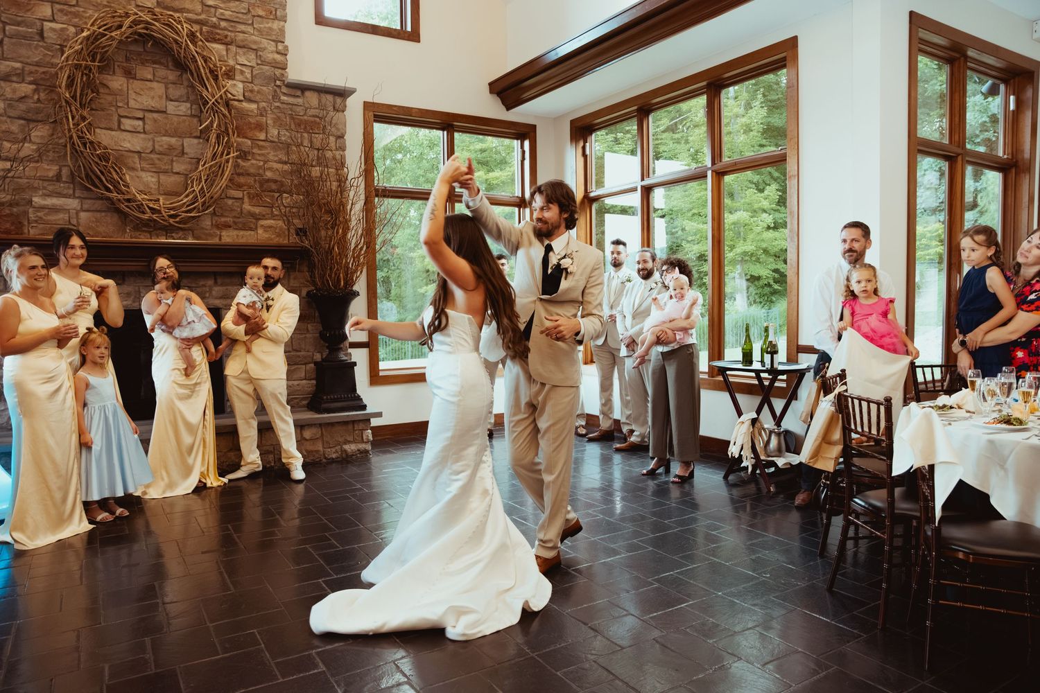 Newlyweds share their first dance as guests applaud in a rustic reception hall with large windows and stone fireplace.
