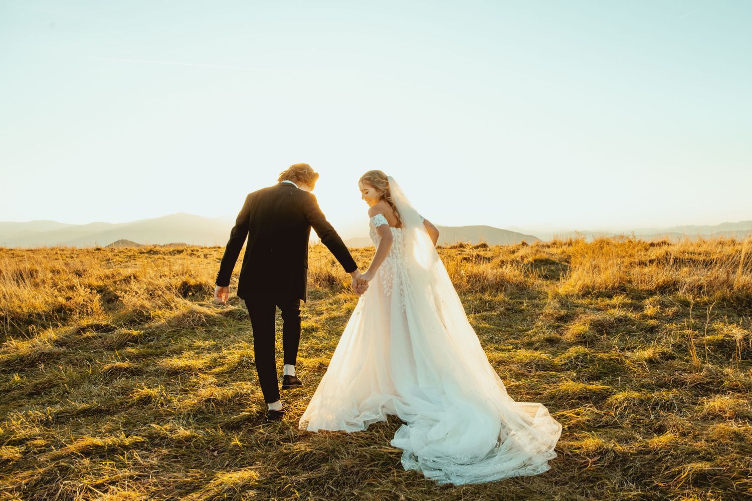 A couple walks through a golden field at sunset in their wedding attire.