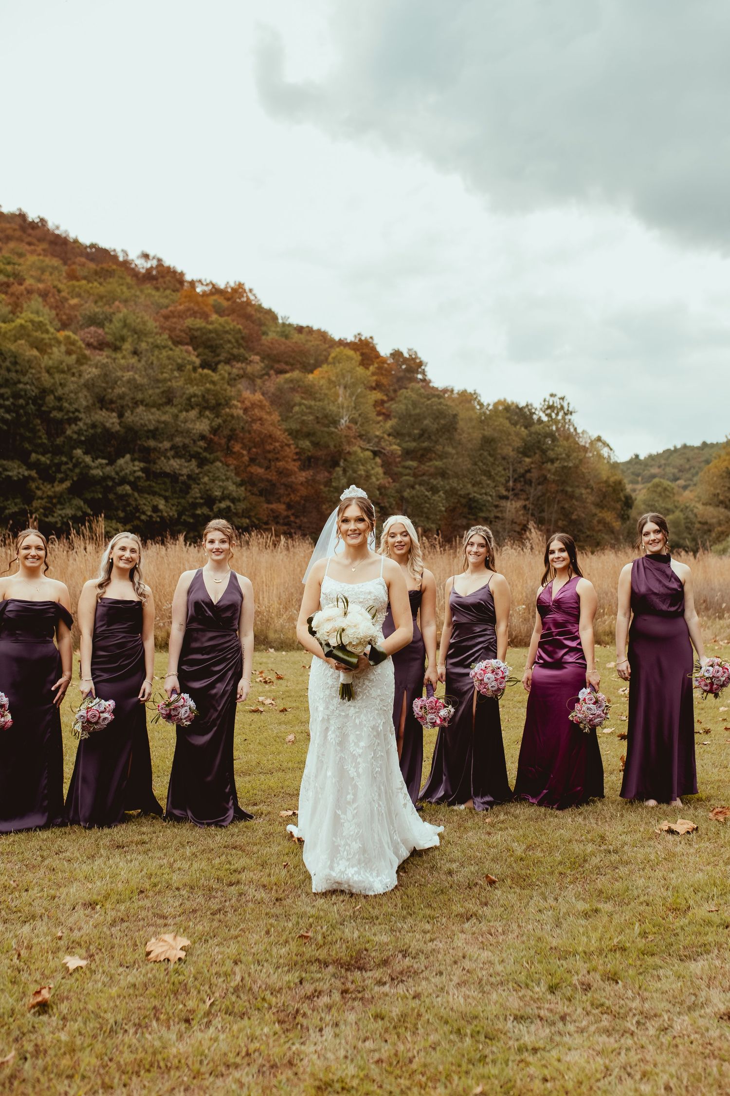 Wedding party in purple bridesmaid dresses poses together in a rustic autumn field during golden hour.