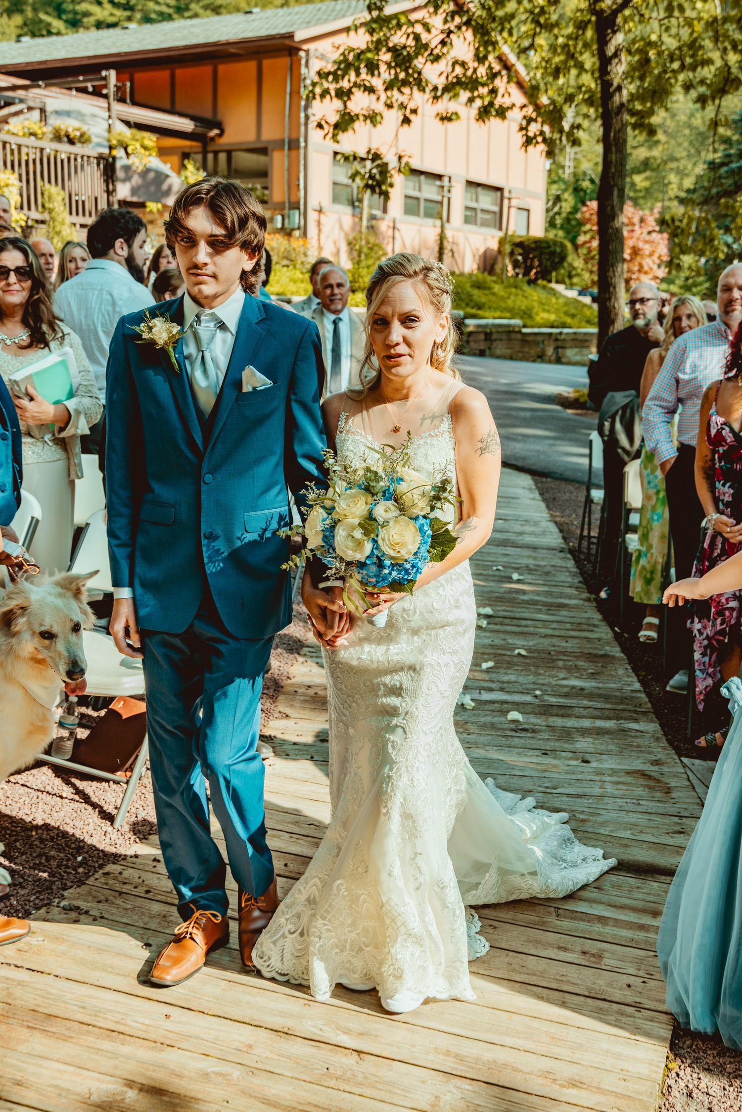 Wedding party in blue suits and white dresses walk along a wooden deck during an outdoor summer celebration.