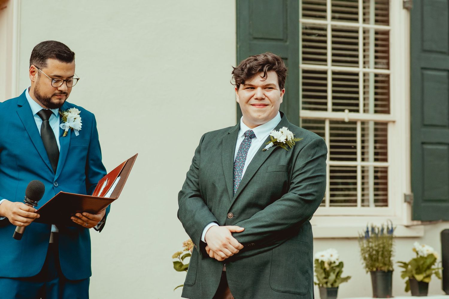 Two people stand together during an outdoor wedding ceremony in front of a white house with green shutters.