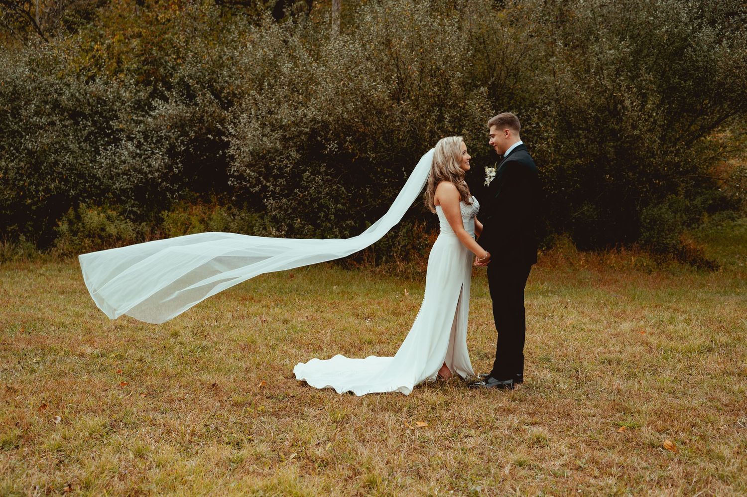 Bride and groom standing in a field, her long veil flowing in the wind, surrounded by autumn trees.
