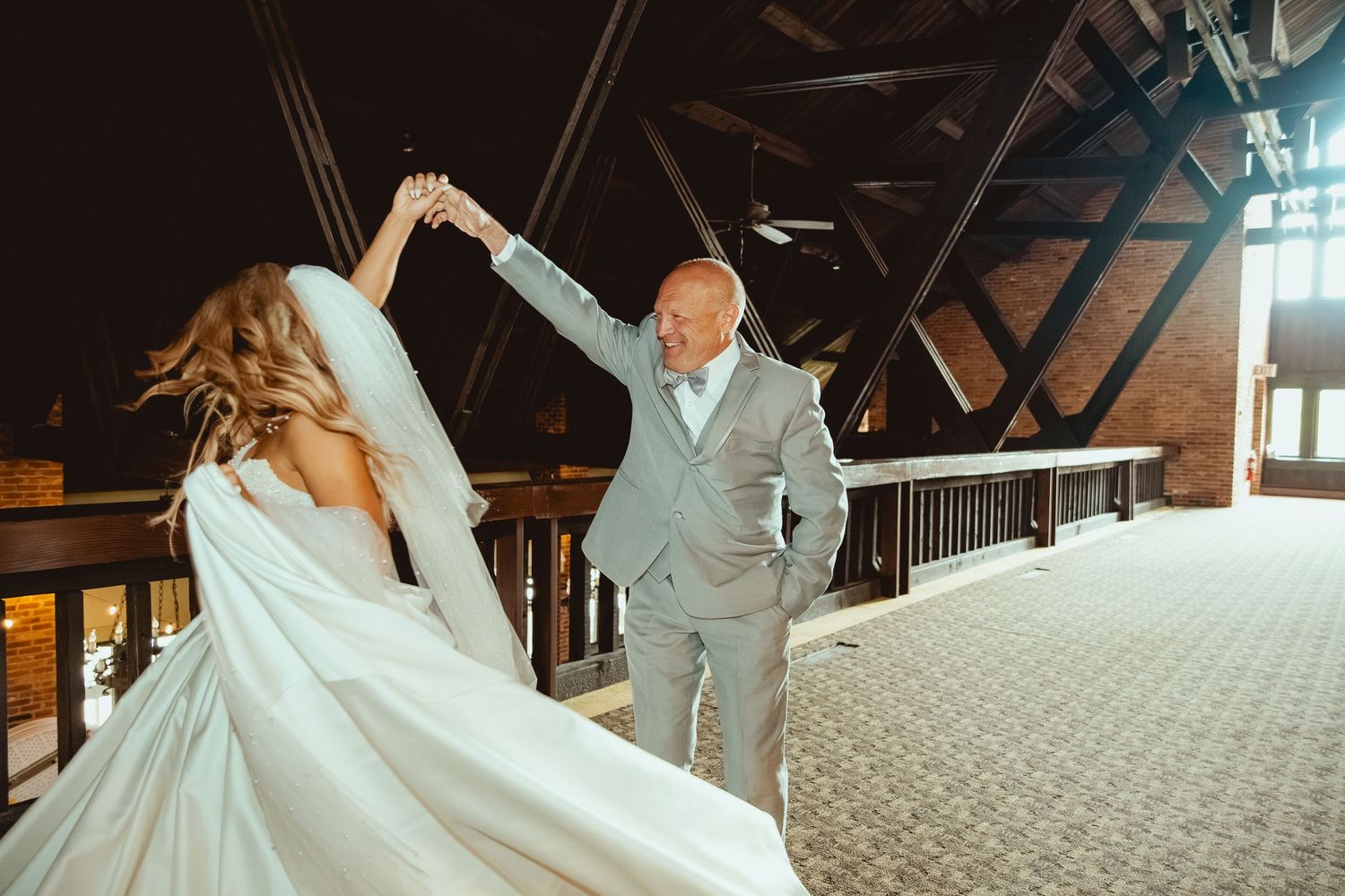 Groom twirling bride in white wedding gown on elegant indoor balcony with wooden beams.