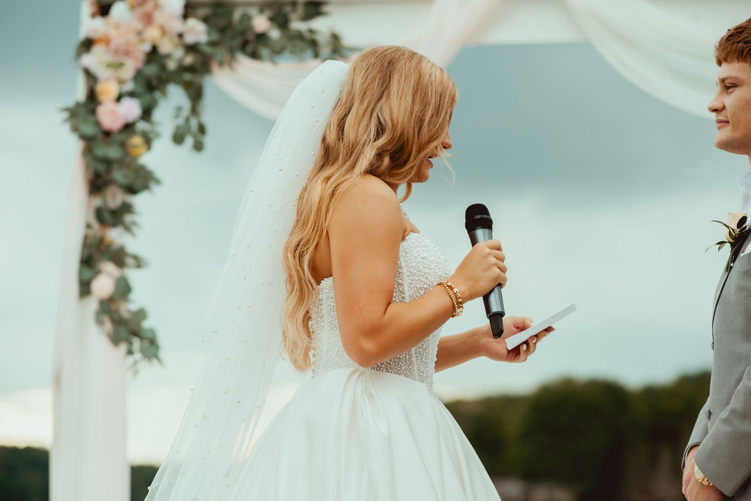 Bride in white gown reading vows at outdoor wedding ceremony with floral arch backdrop.
