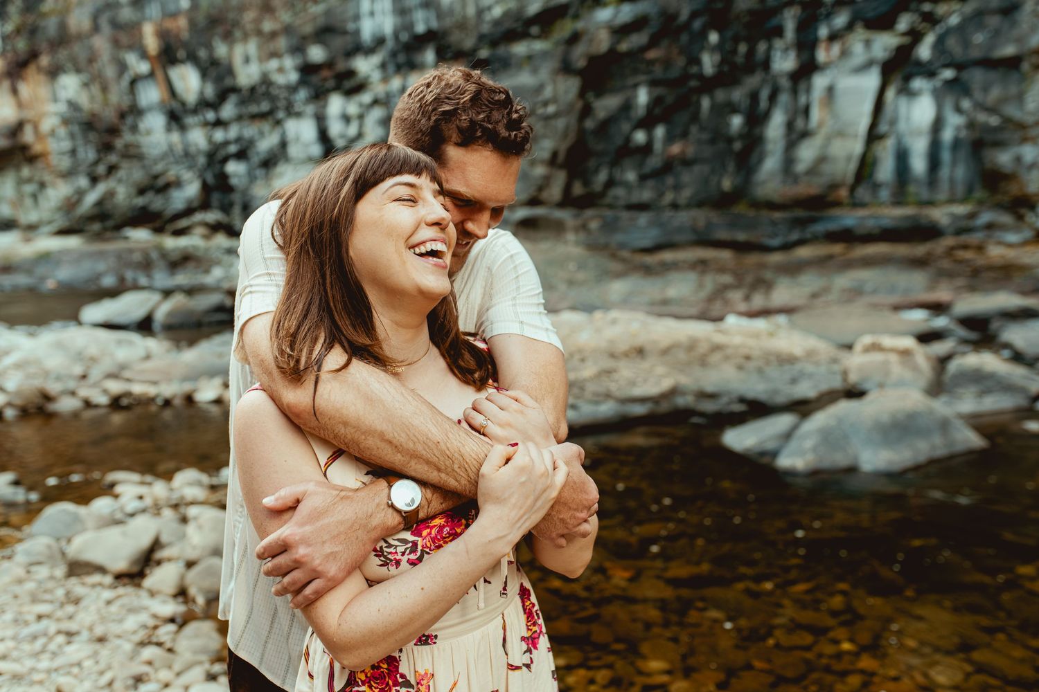 Couple embracing and laughing by a rocky river, woman in floral dress, man in white shirt, outdoor engagement photo.