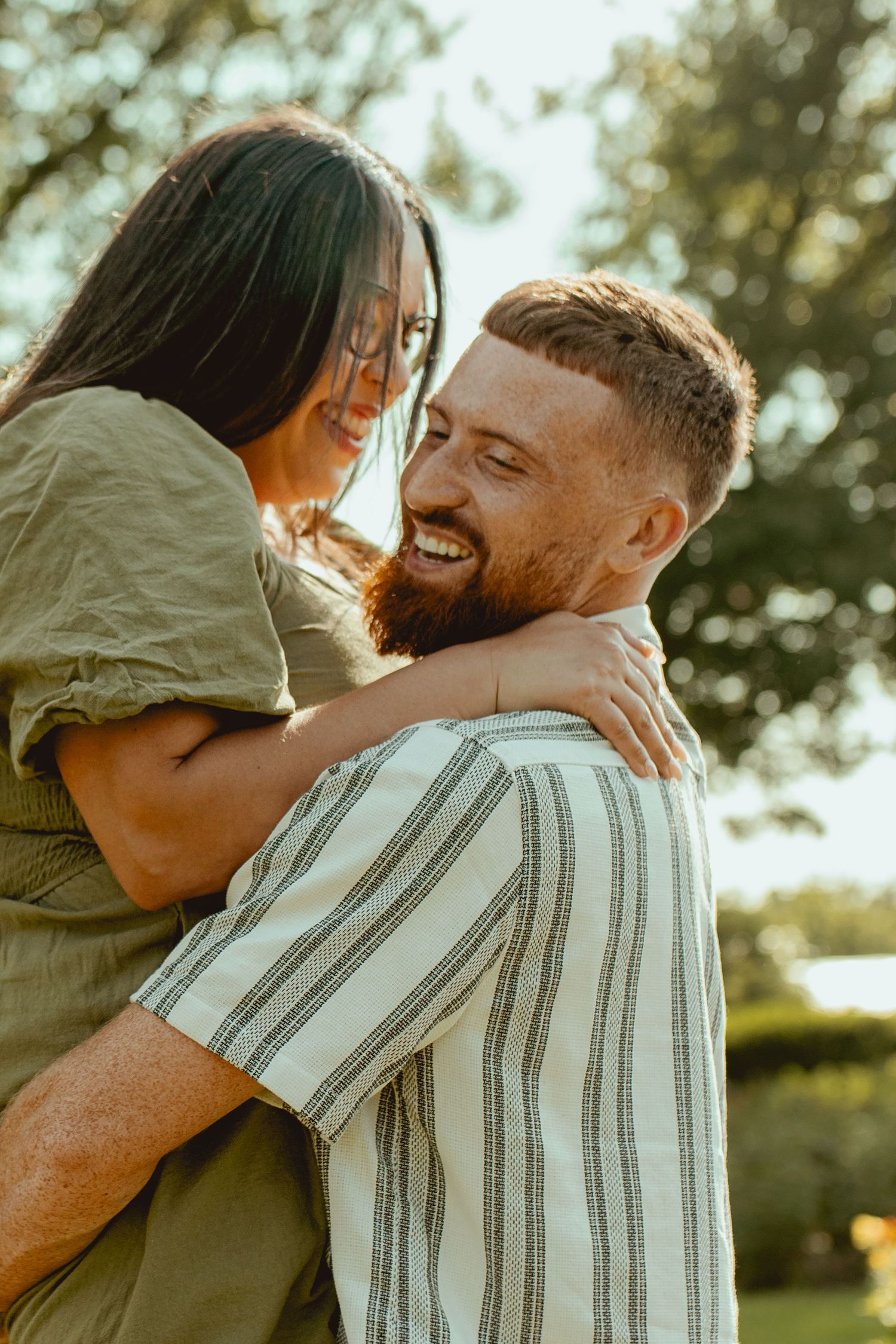 A smiling bearded man lifts a laughing woman in a sunny park, both wearing casual green and striped outfits.
