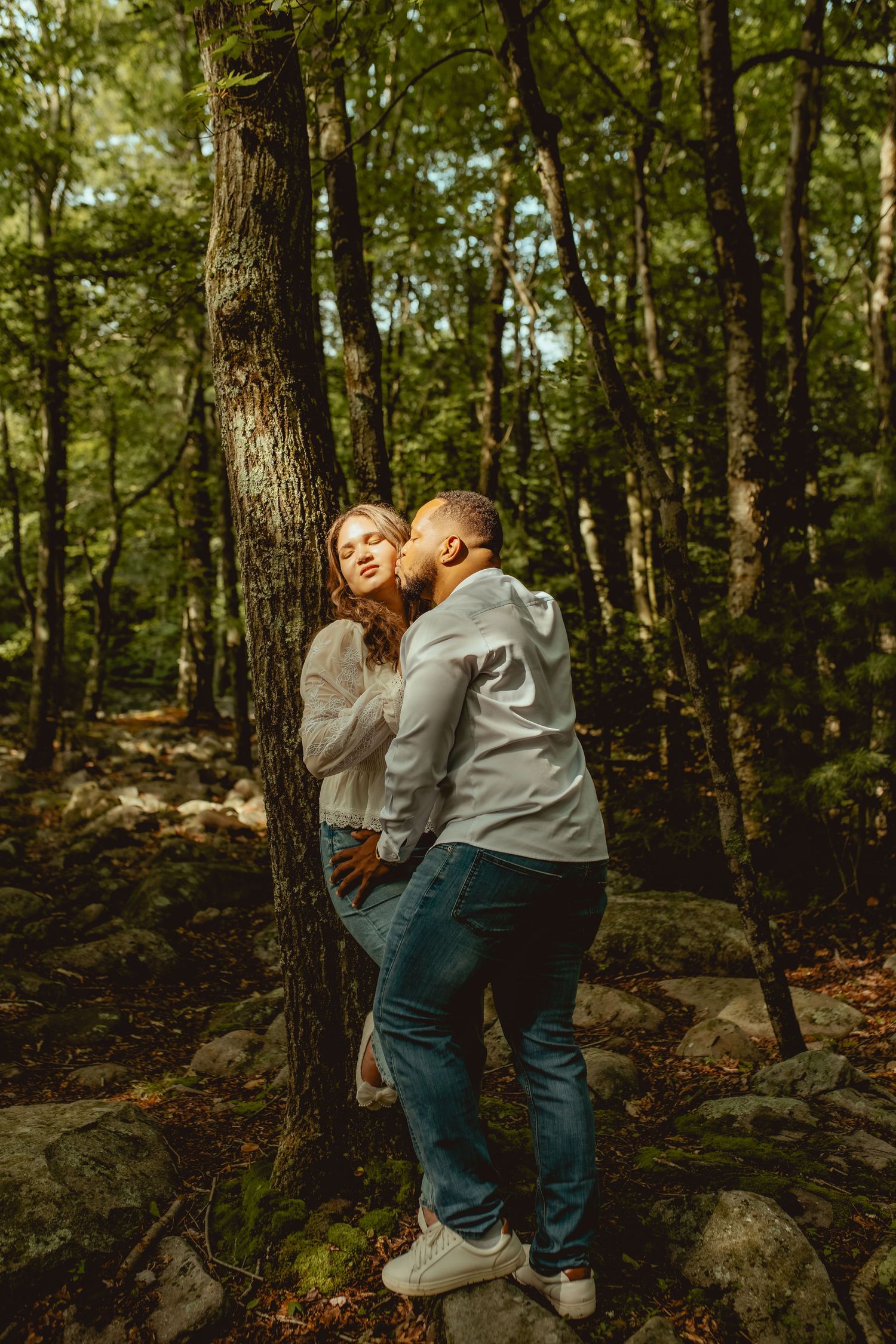 A romantic couple shares a kiss leaning against a tree in a lush green forest setting.