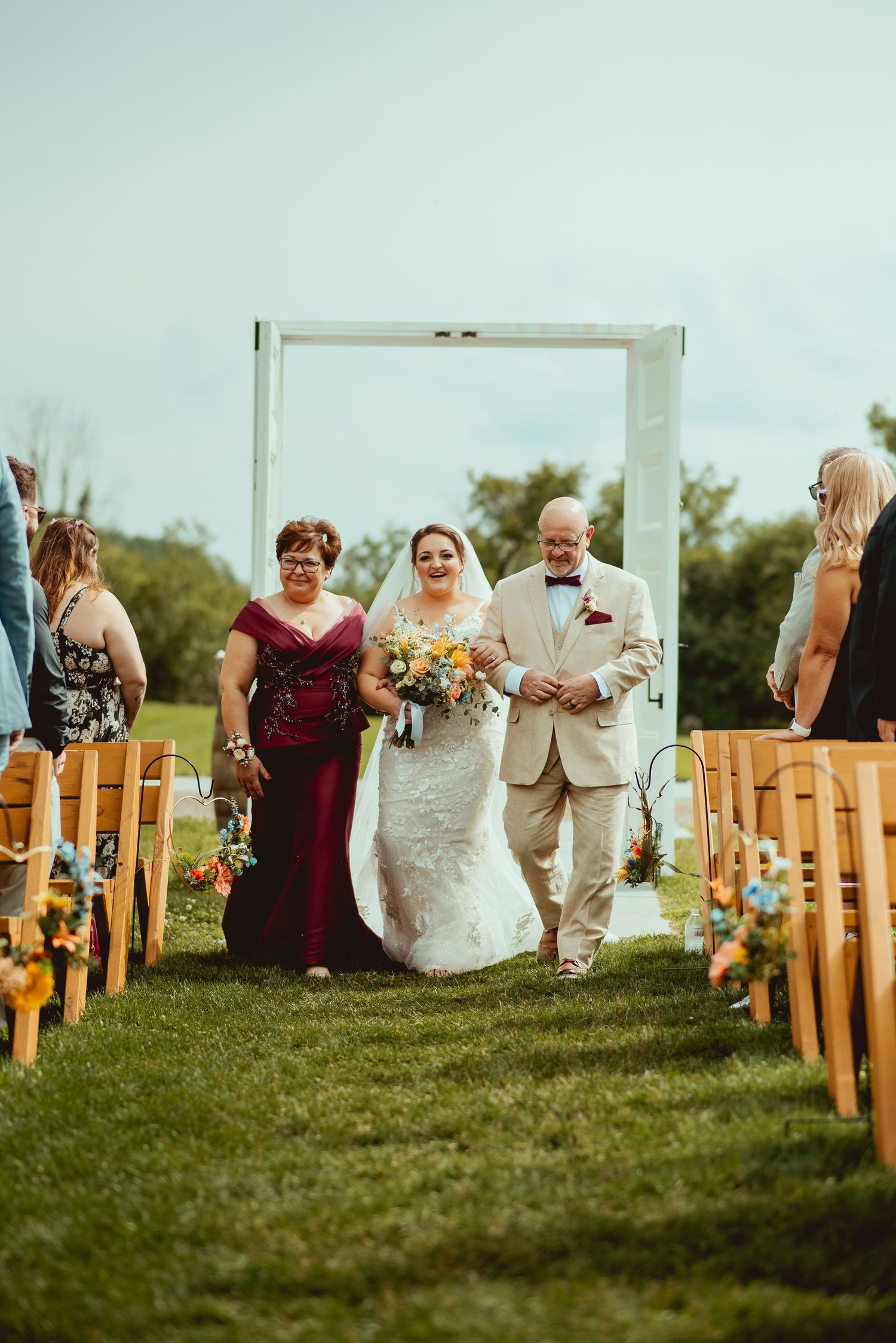Bride walks down outdoor aisle flanked by parents, wearing lace gown, holding colorful bouquet at sunny garden wedding ceremony.