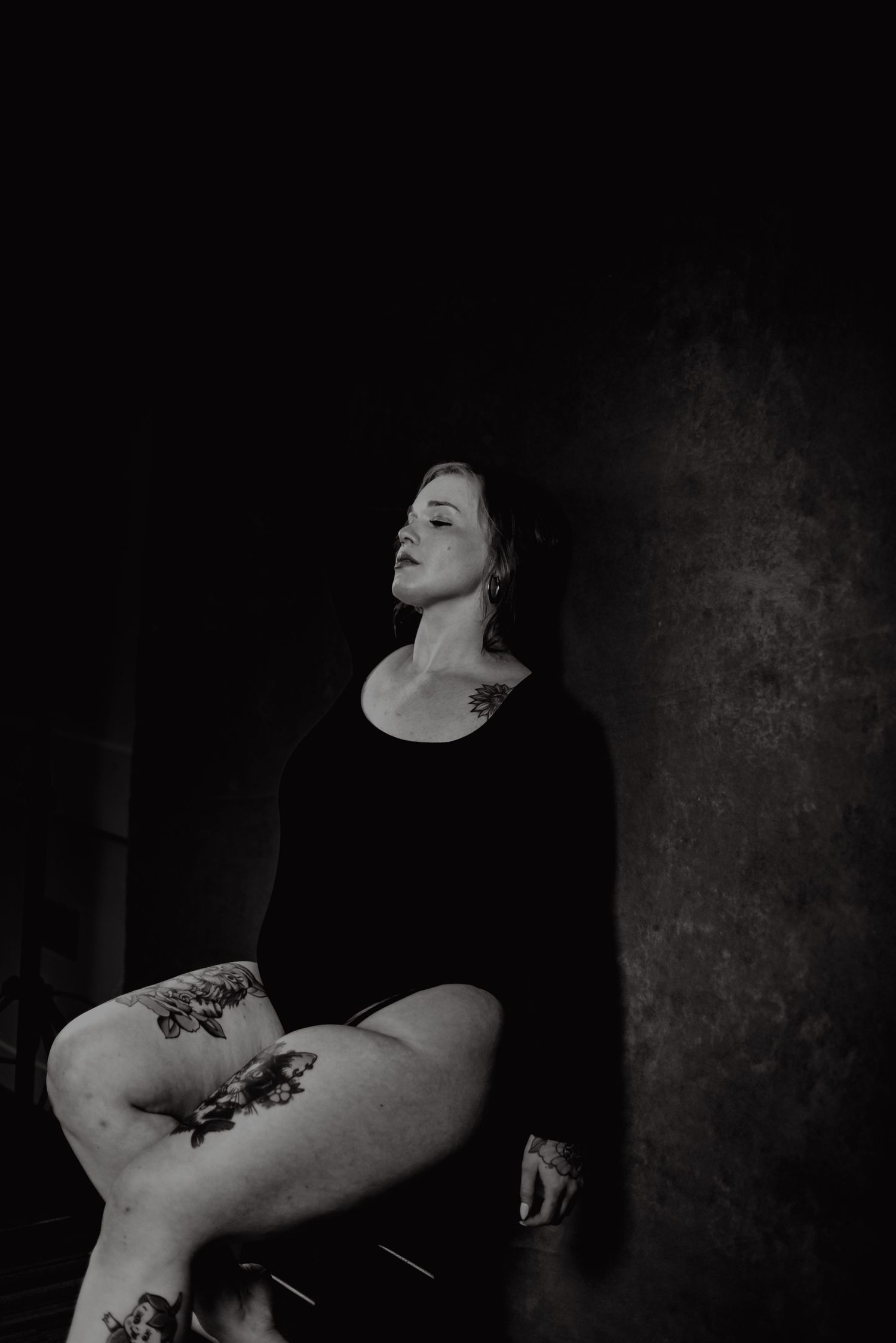 Black and white photo of a tattooed woman in a black dress sitting confidently in dramatic moody lighting.