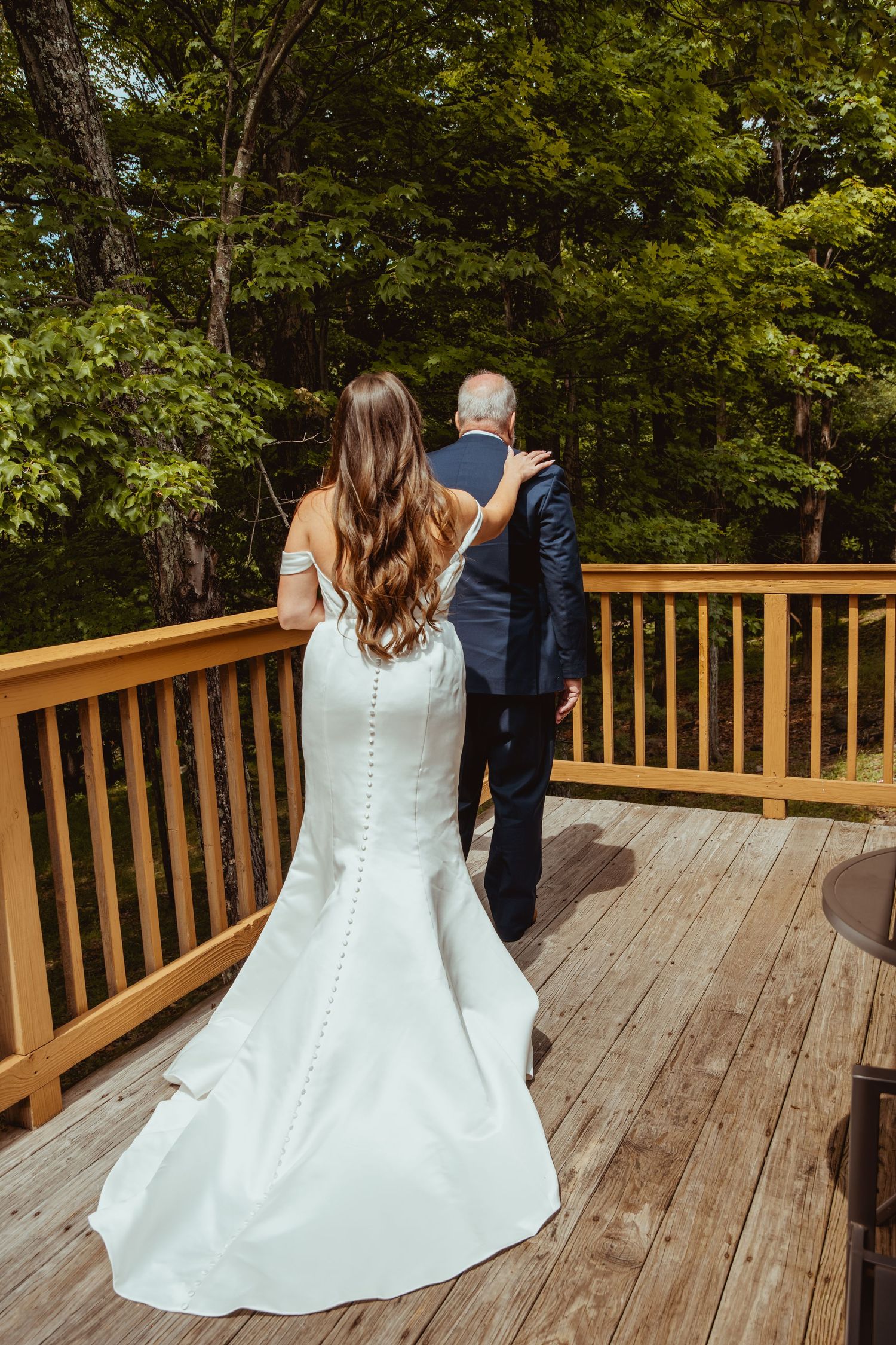 Bride in white gown with button back stands with older man in navy suit on wooden deck surrounded by lush green trees.