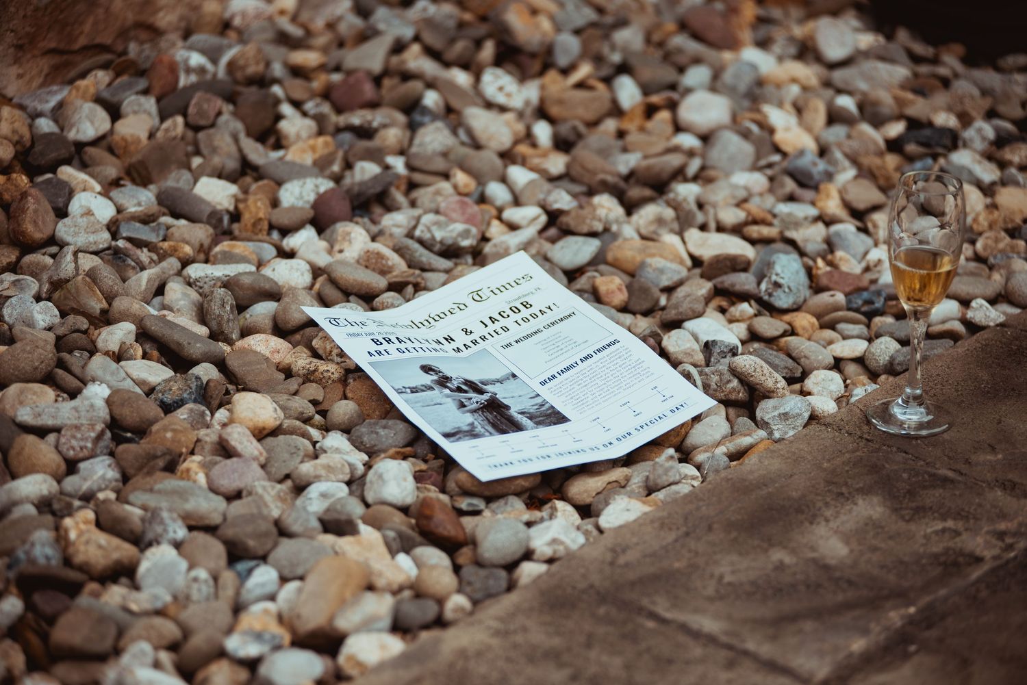 Vintage newspaper and champagne glass rest on decorative pebble surface at outdoor wedding reception.