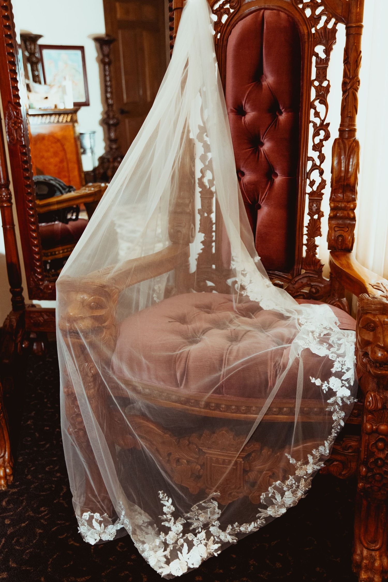 Elegant lace-trimmed wedding veil draped over ornate tufted burgundy throne chair indoors.