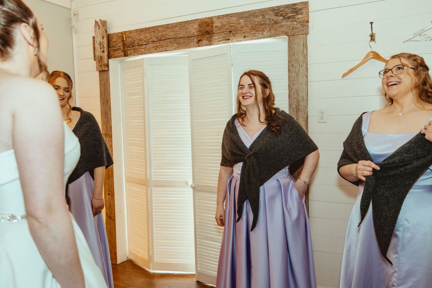 Bridesmaids in lavender gowns and dark shawls laughing together in rustic wedding preparation room.