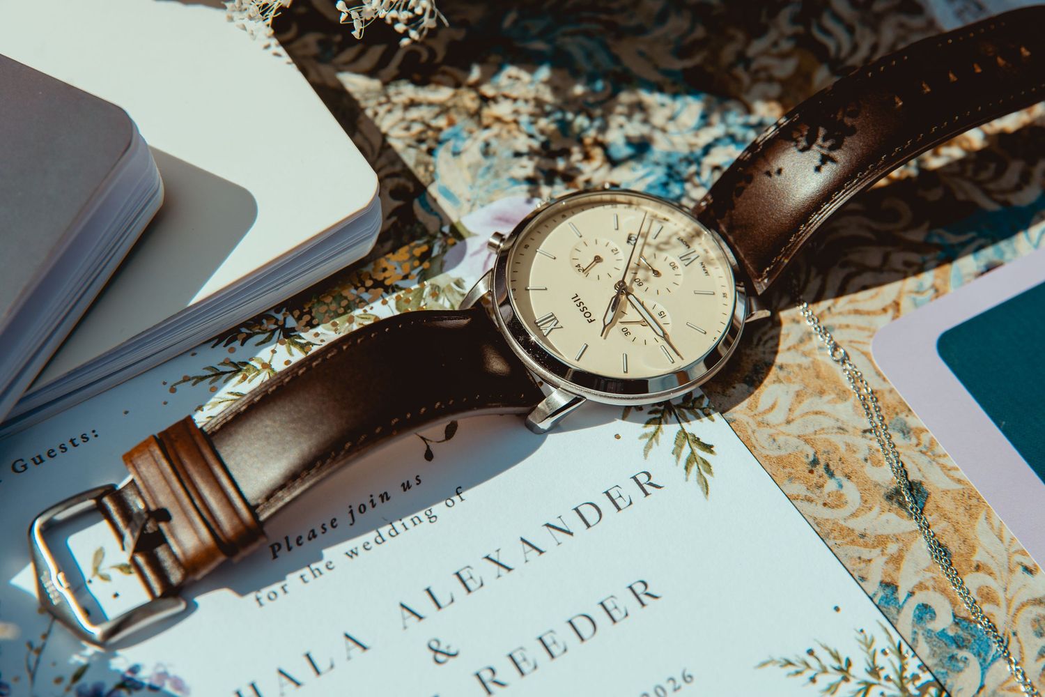 Elegant brown leather wristwatch with cream dial resting on floral wedding invitation cards.