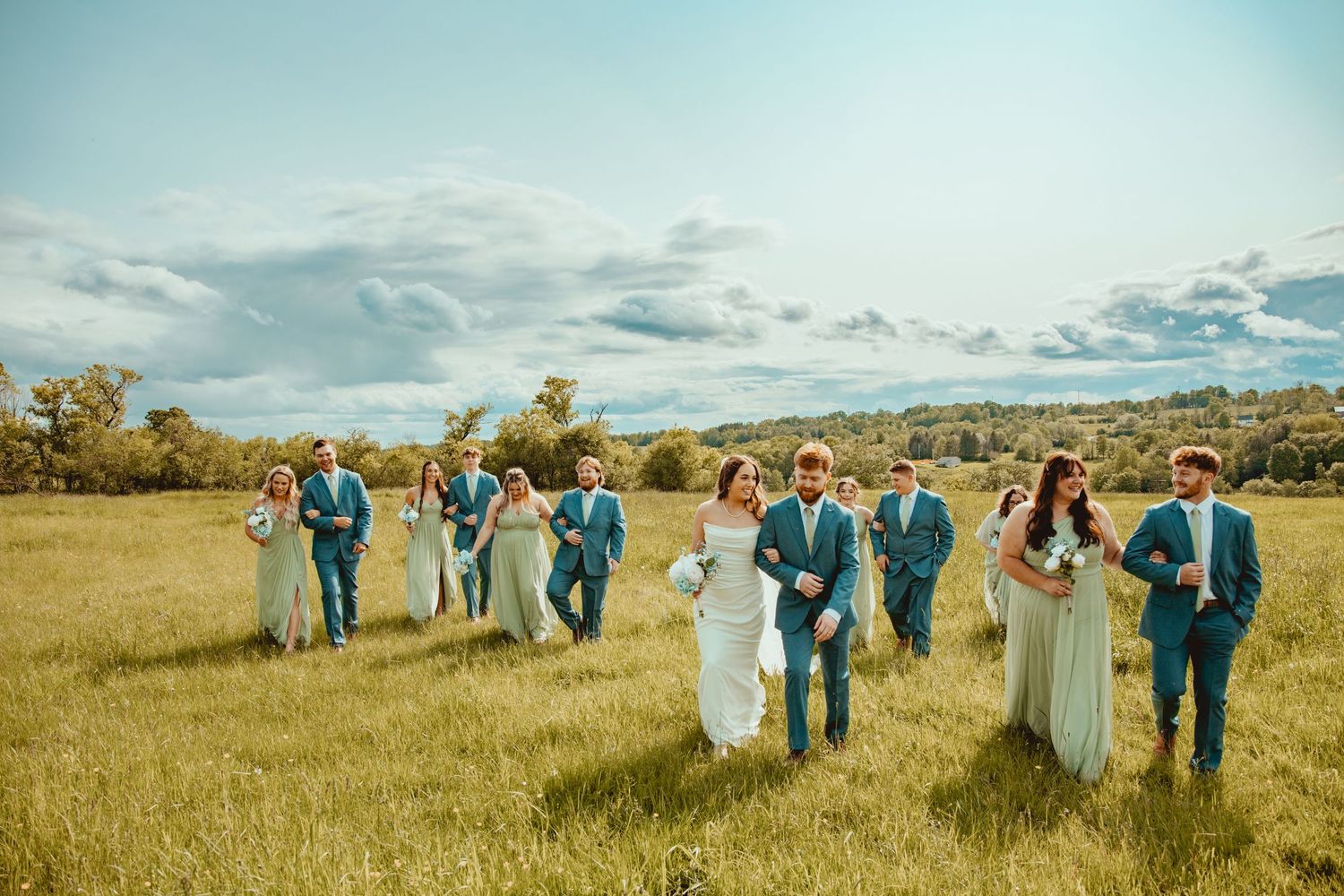 Wedding party walking through a golden field under blue skies wearing blue suits and mint bridesmaid dresses.
