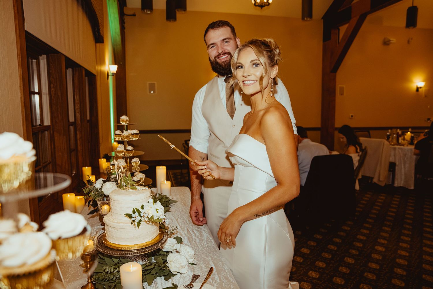 A couple cuts their elegant white wedding cake at an indoor reception venue with warm lighting.