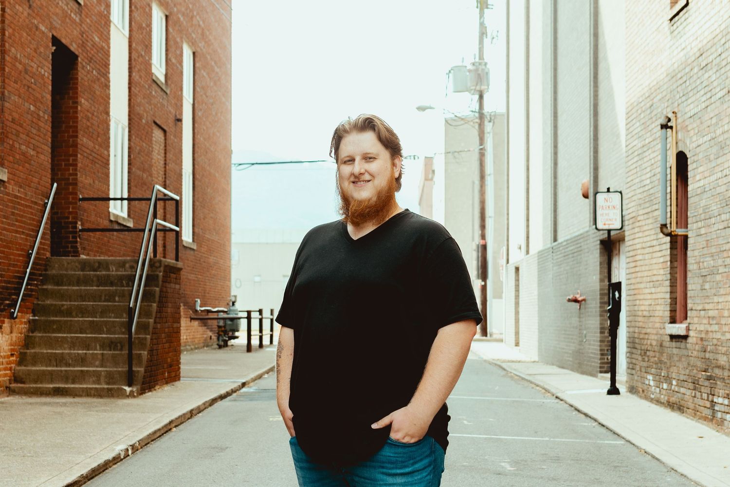 A person stands against a white wall in a minimalist urban setting wearing dark clothing.