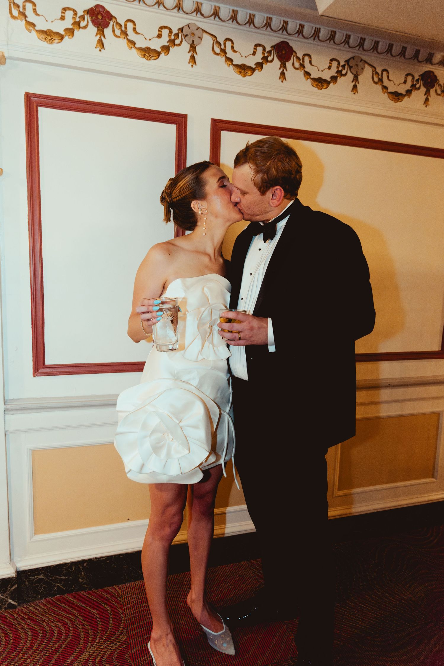 Couple dancing in formal attire beneath ornate crown molding and decorated walls.