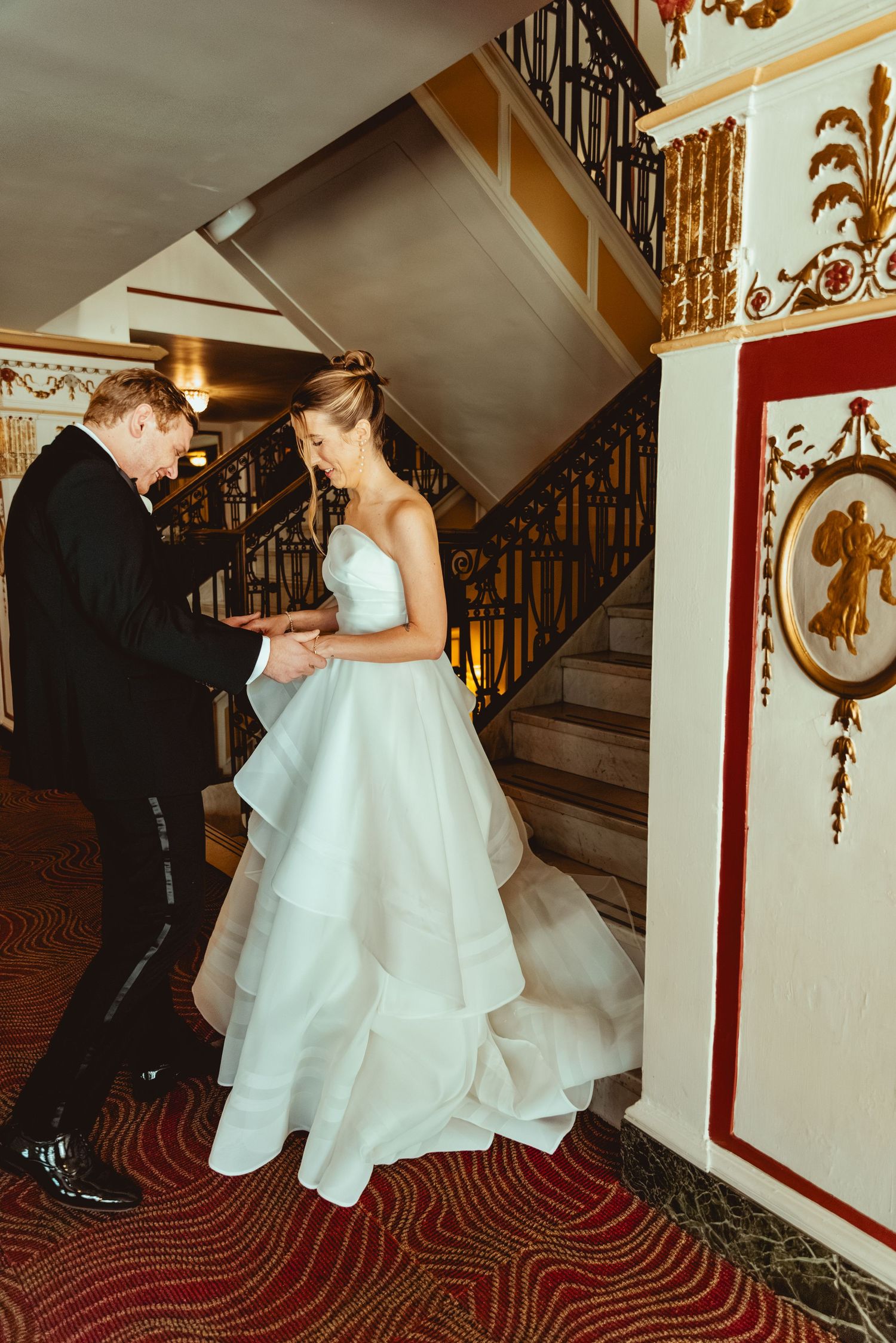 Wedding couple descending an elegant staircase with red carpeting and ornate gold trim details.