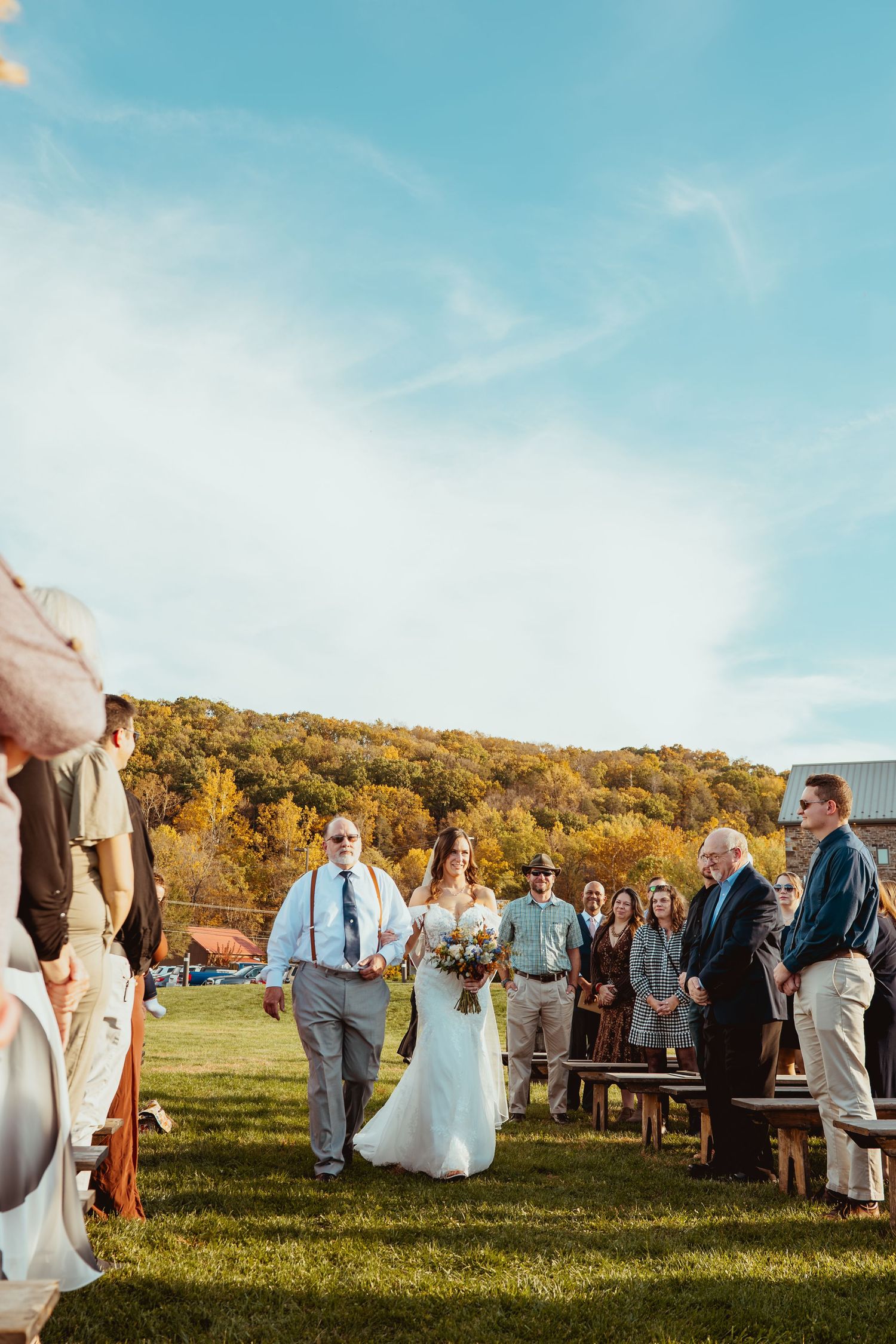 Wedding guests gather in a sunny field during golden hour with rolling hills in the background.