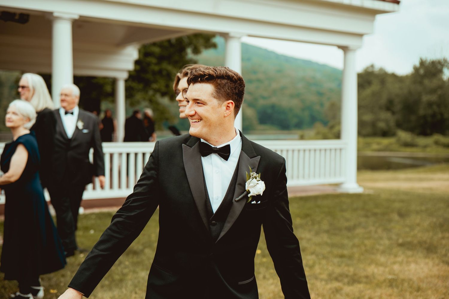 Groomsmen in black suits laughing together on a sunny day at an outdoor wedding venue with rolling hills in background.