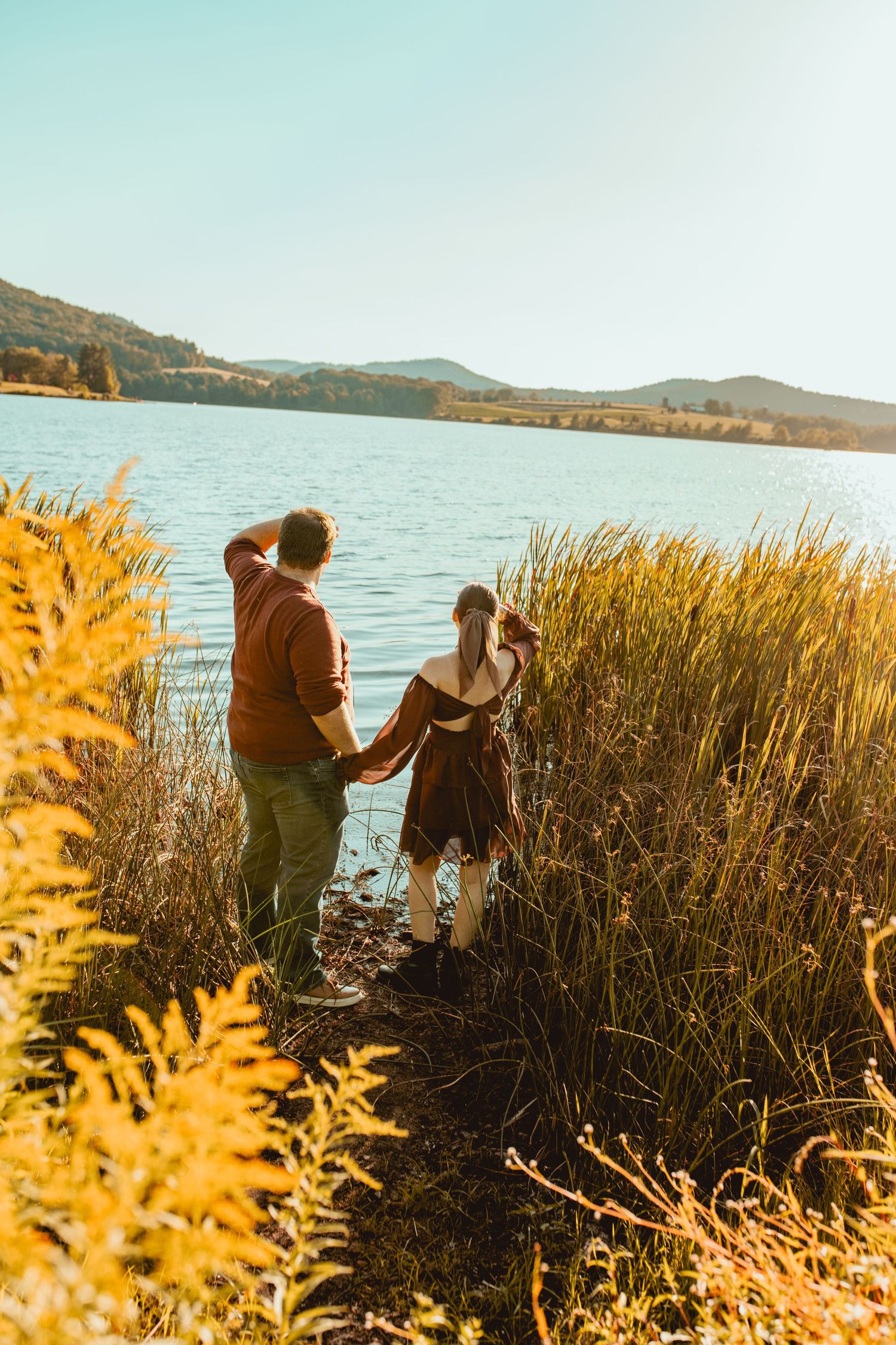 People explore a scenic lakeside with tall golden grasses at sunset, mountains visible in the background.
