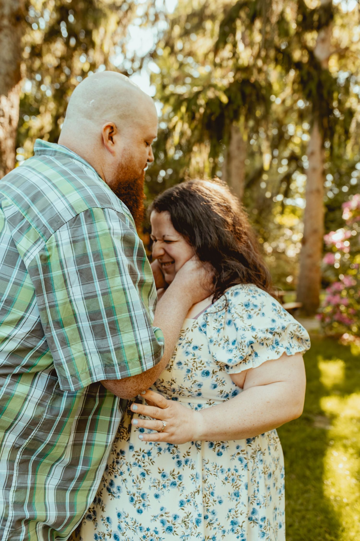 A couple shares a tender moment outdoors, the man in plaid holding the laughing woman in a floral dress among trees.