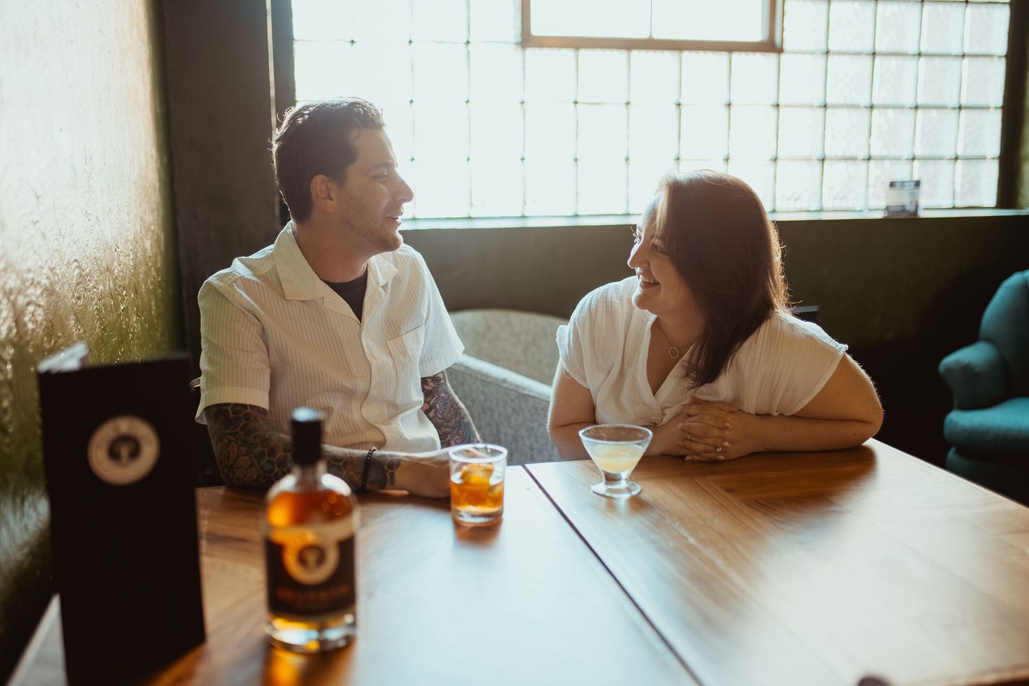 Couple smiling and enjoying cocktails and whiskey at a cozy bar table in a warmly lit restaurant.
