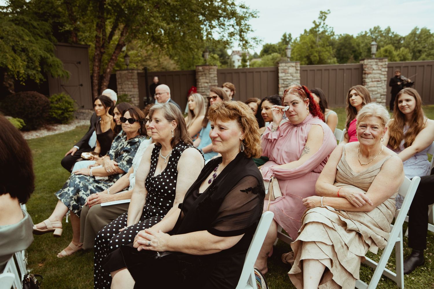 Guests seated in white chairs at an outdoor wedding ceremony on a sunny day in a garden setting.