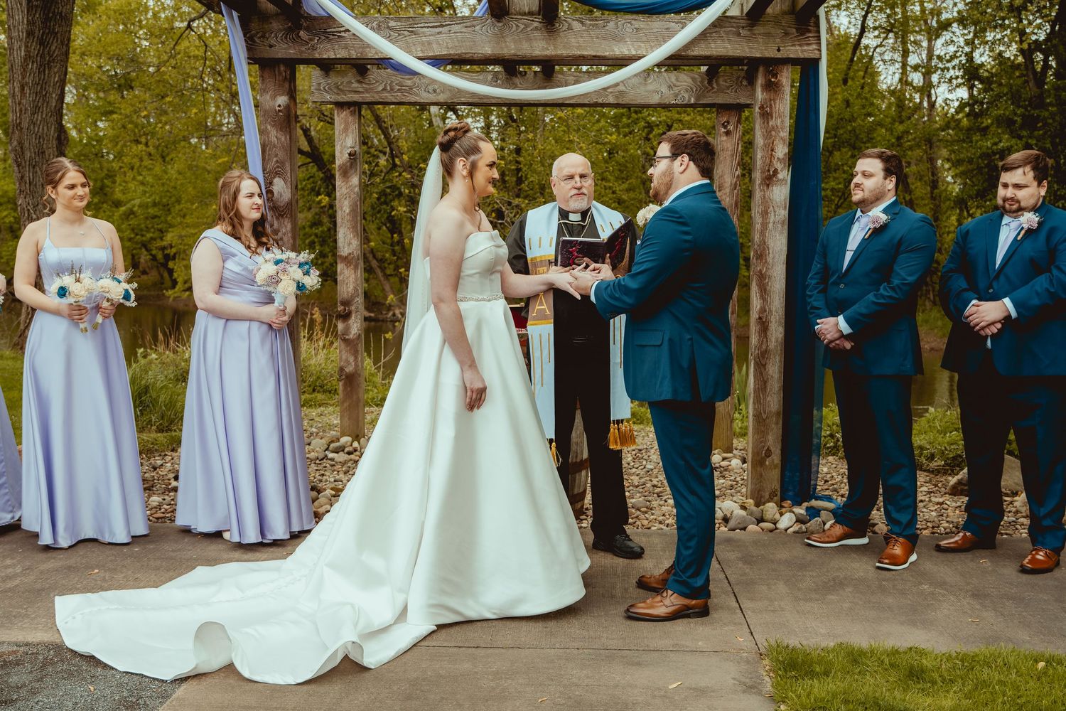 Bride and groom exchanging vows at outdoor wedding ceremony with bridesmaids in lavender and groomsmen in navy.
