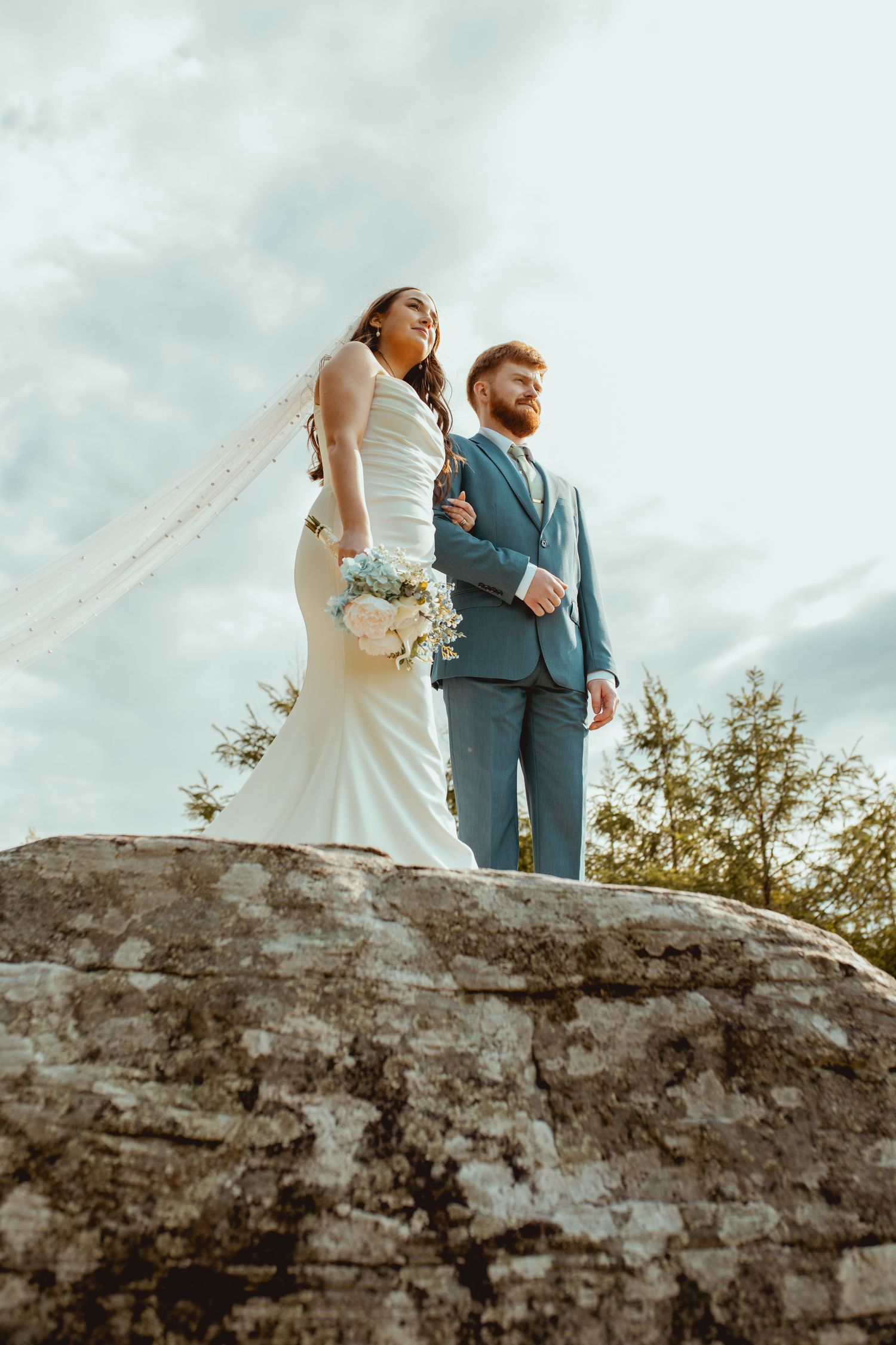Couple in wedding attire standing atop rocky outcrop against cloudy sky.