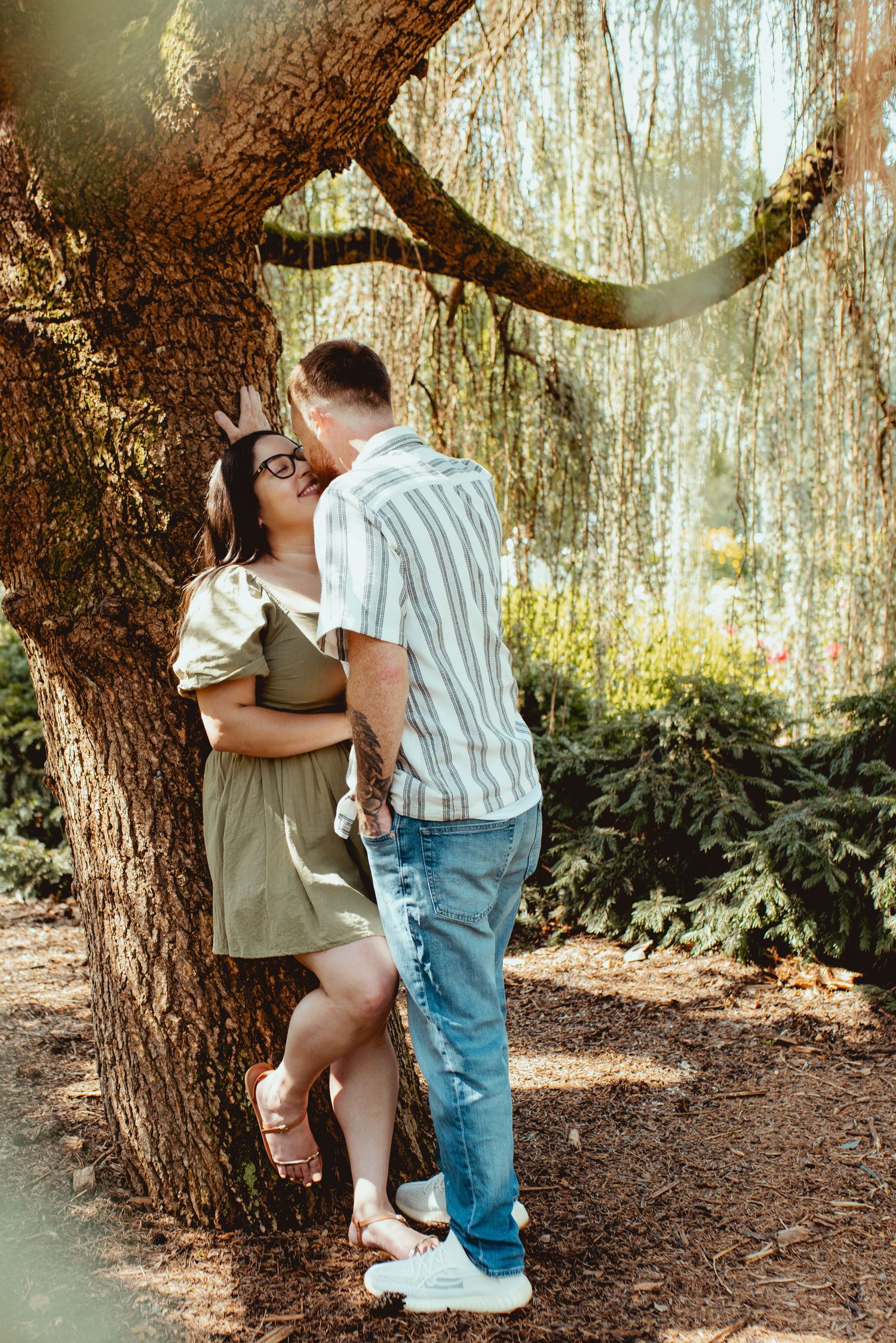 Couple sharing a romantic kiss under a weeping willow tree in a lush garden setting.
