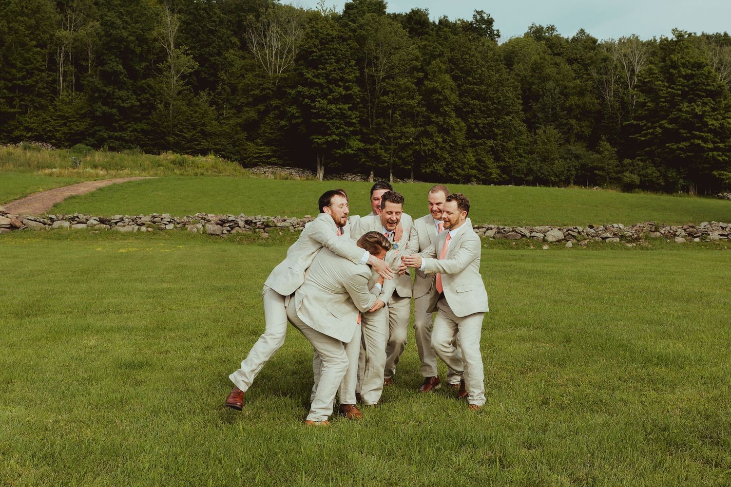 Groomsmen in light suits laughing and hugging on green lawn with stone wall and forest backdrop.